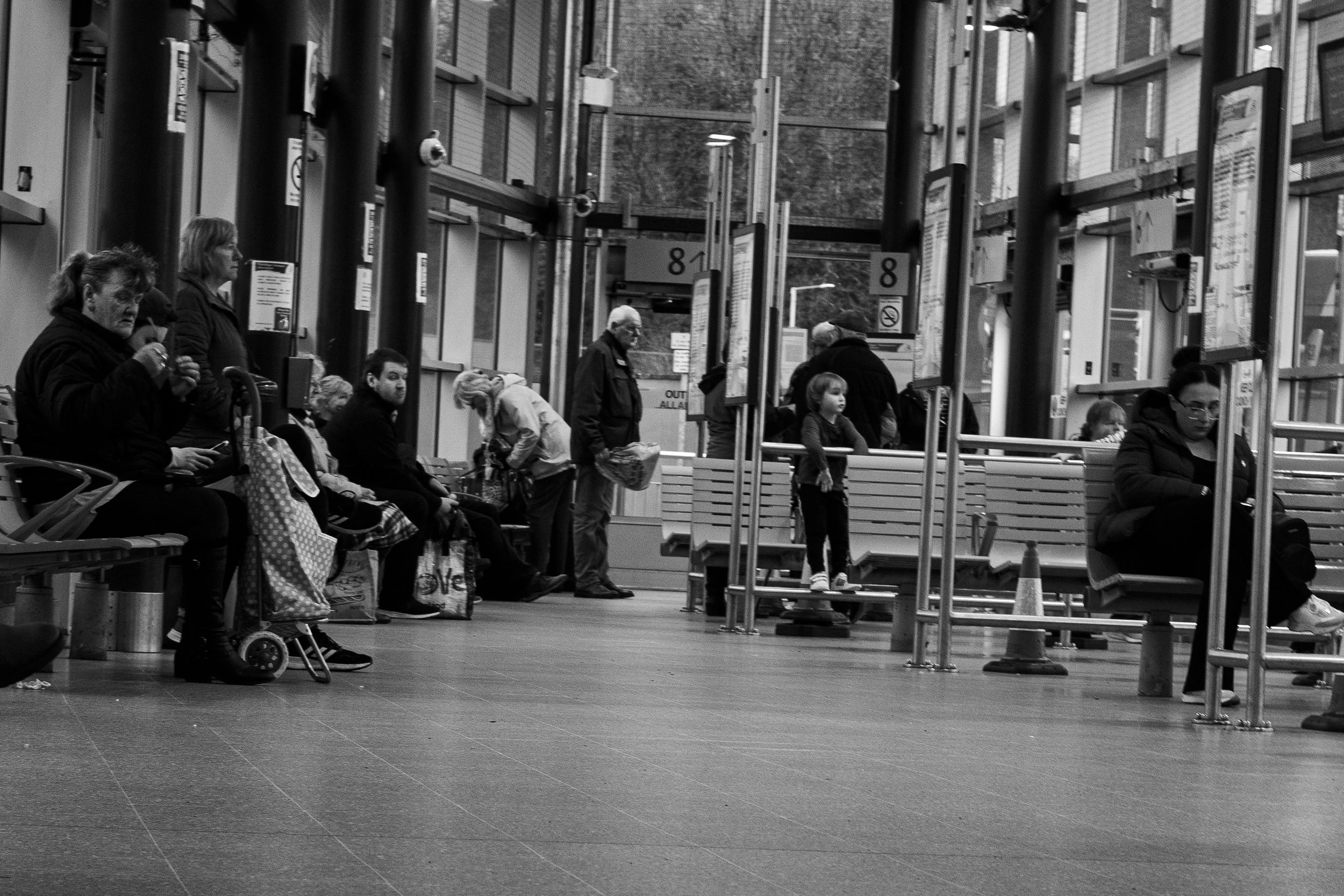 People waiting inside a public transportation station, sitting on benches and standing, with some using phones or carrying bags, in a spacious, well-lit area with large windows.