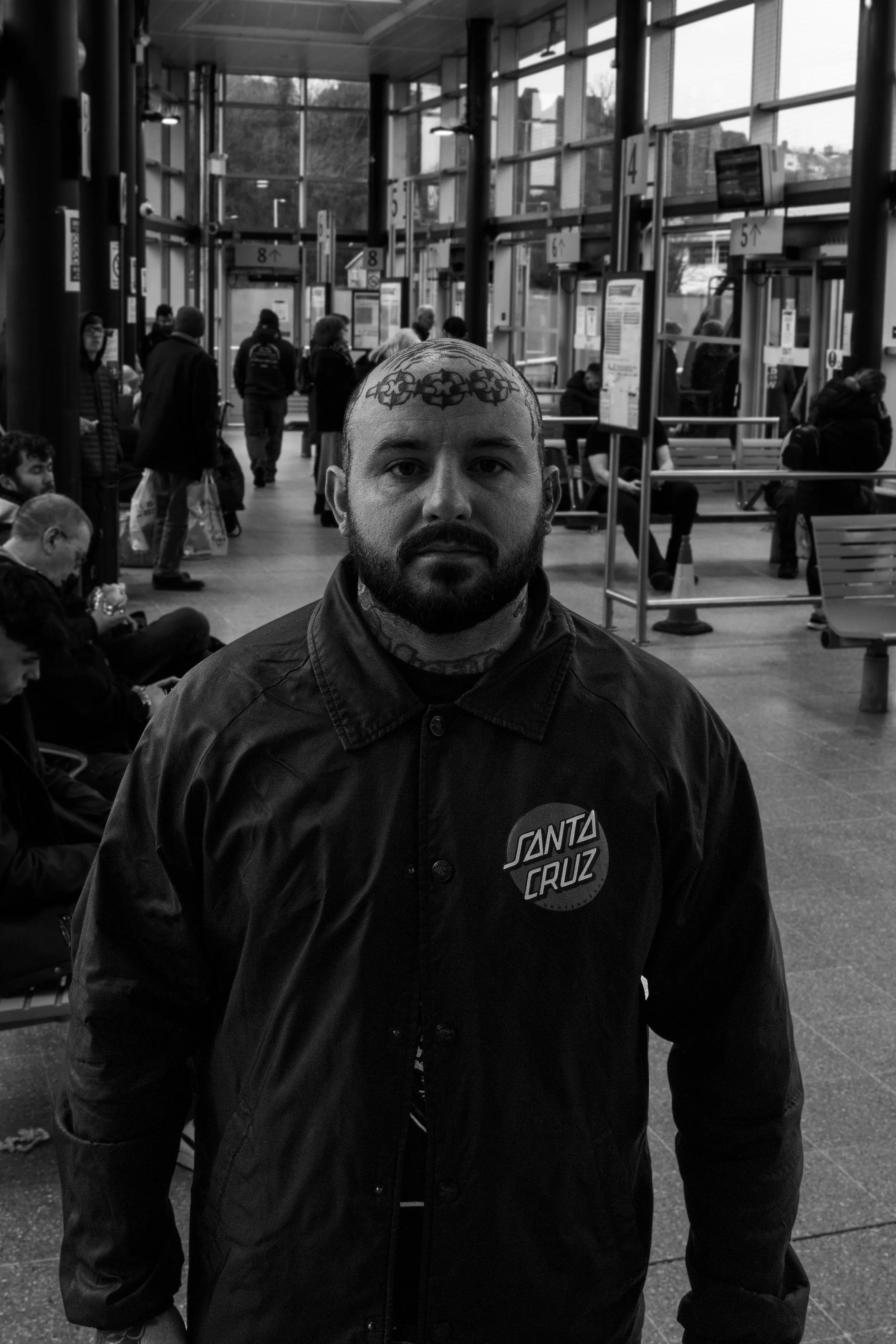 A man with tattoos on his head and neck, wearing a Santa Cruz jacket, stands in a busy transportation hub with other people waiting and walking in the background.