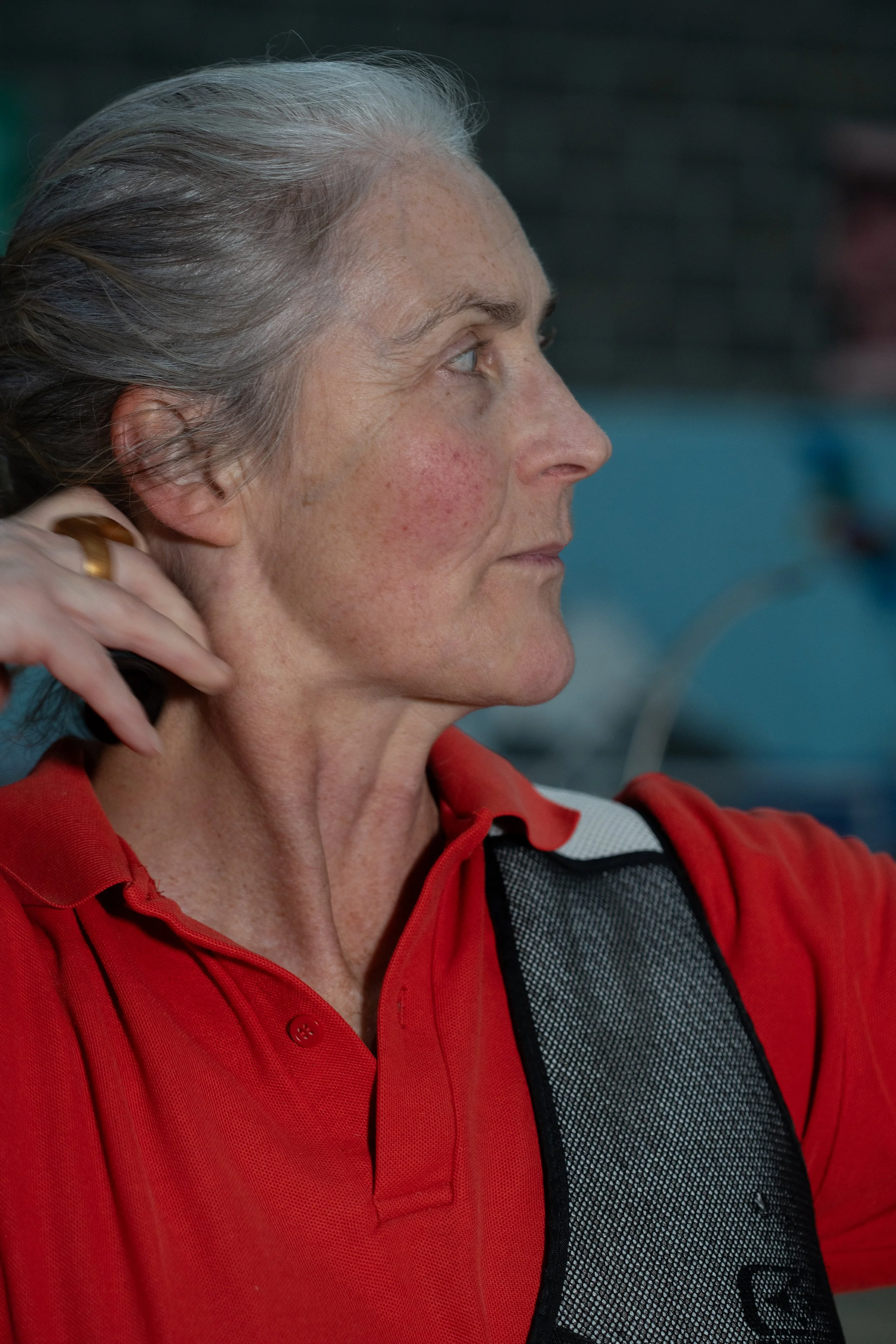 Close-up profile of an older woman with gray hair, wearing a red polo shirt and a black vest, with her hand touching her neck.