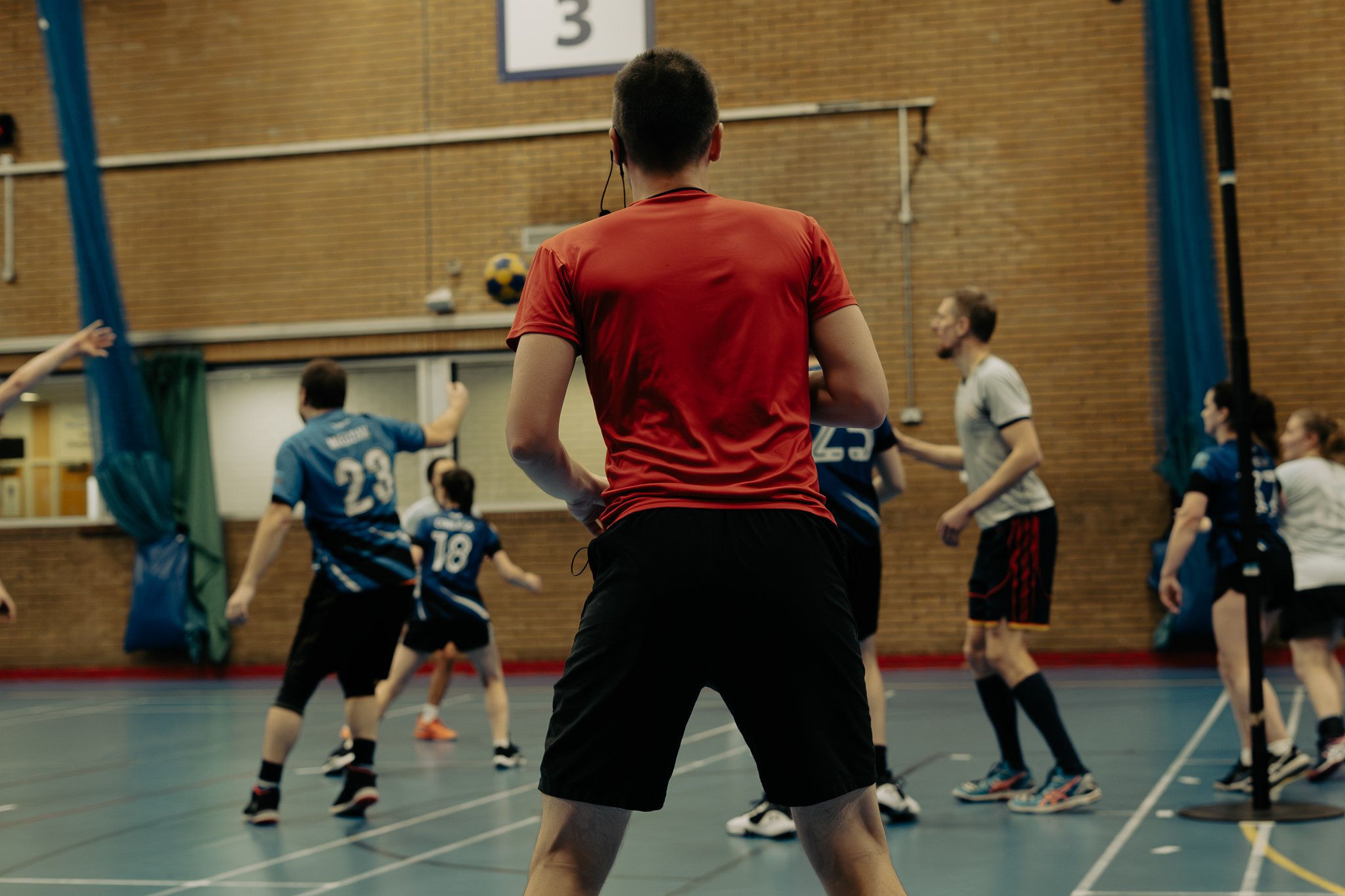 Indoor sports scene with a coach or referee in a red shirt overseeing a game with players in blue and white jerseys.