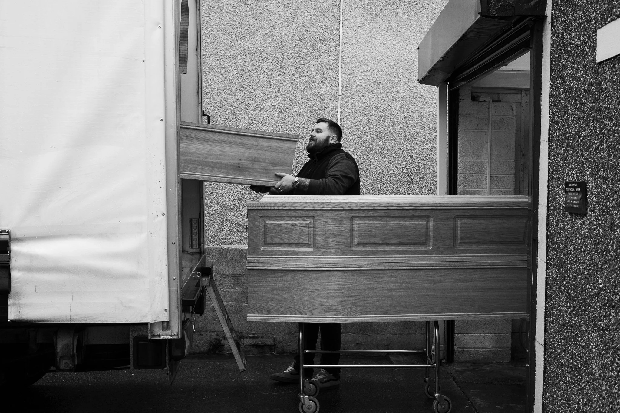 A man with a beard and tattoos wearing a black hoodie is loading a wooden piece of furniture onto a truck. He is standing on a wheeled dolly near a textured wall with a small window.