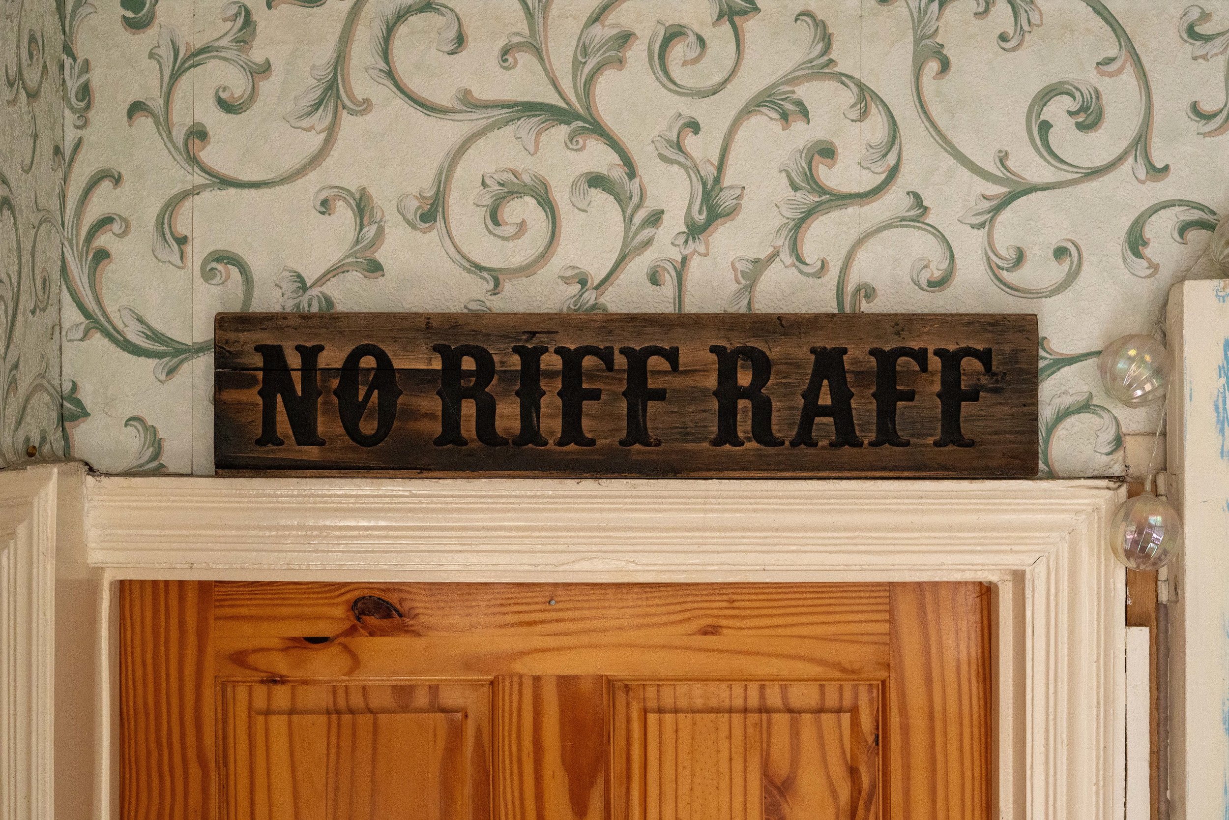 A wooden sign with the words 'NO RIF RAF' in black letters, mounted above a wooden door frame, with floral patterned wallpaper in the background.