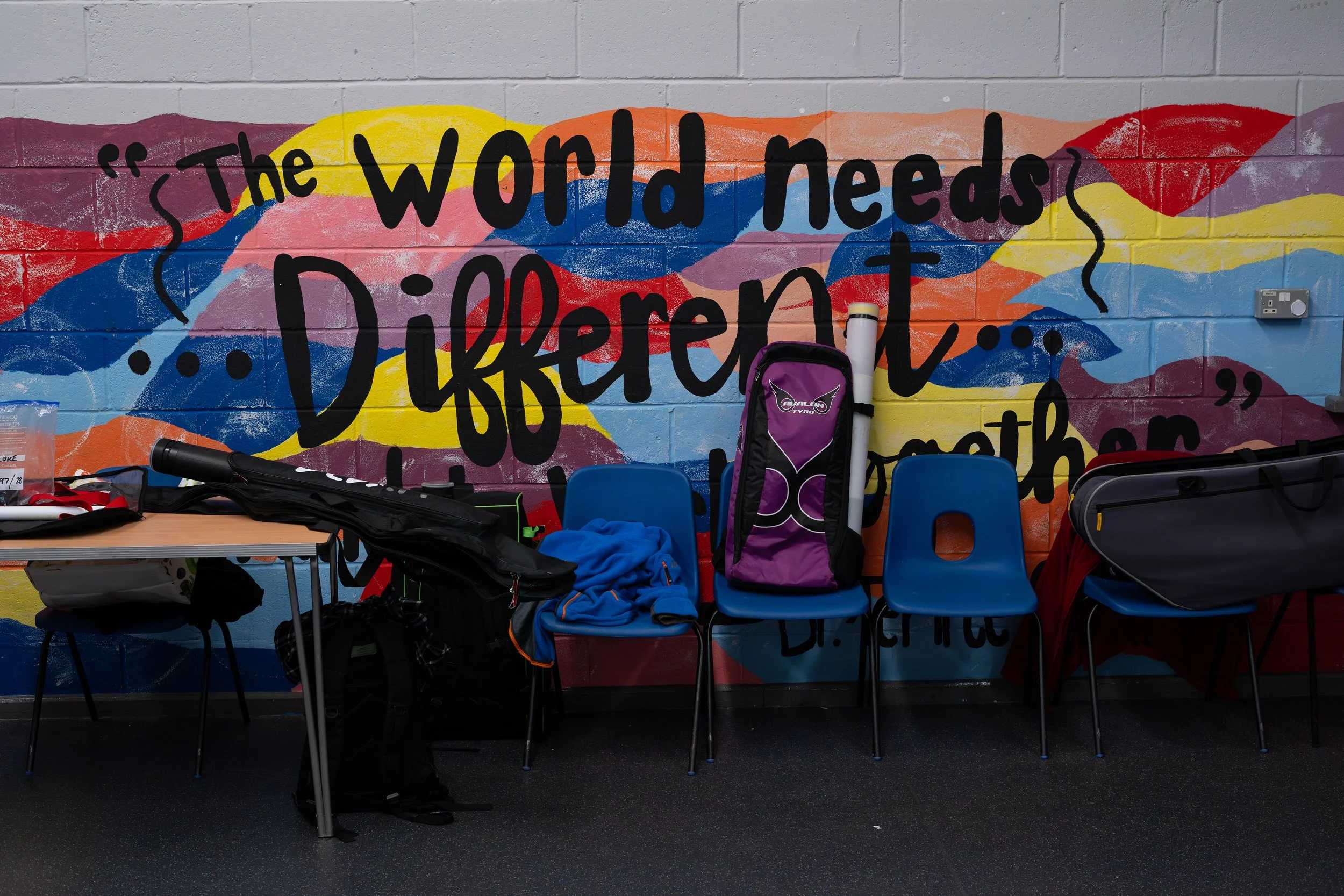 Colorful mural on a brick wall displaying the quote 'The world needs different' with chairs, backpacks, and sports equipment in front of it.