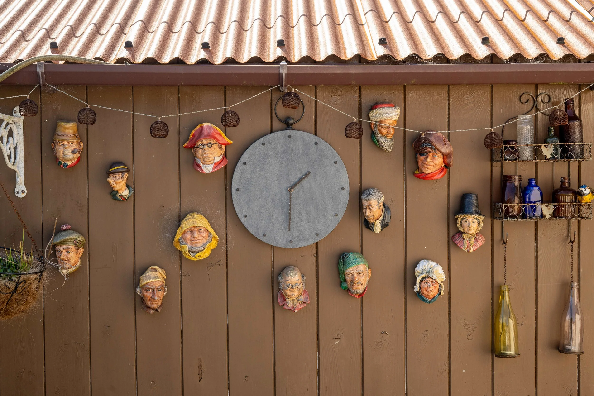 Decorative outdoor wall with a clock and multiple colorful face sculptures, shelves with bottles, and a hanging basket.