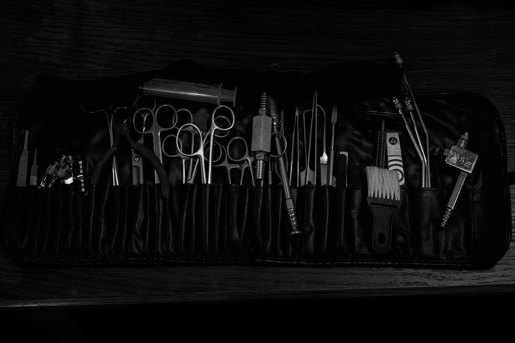 Medical or surgical tools arranged in a black roll-up case on a wooden surface, including scissors, forceps, a brush, syringe, and other instruments.