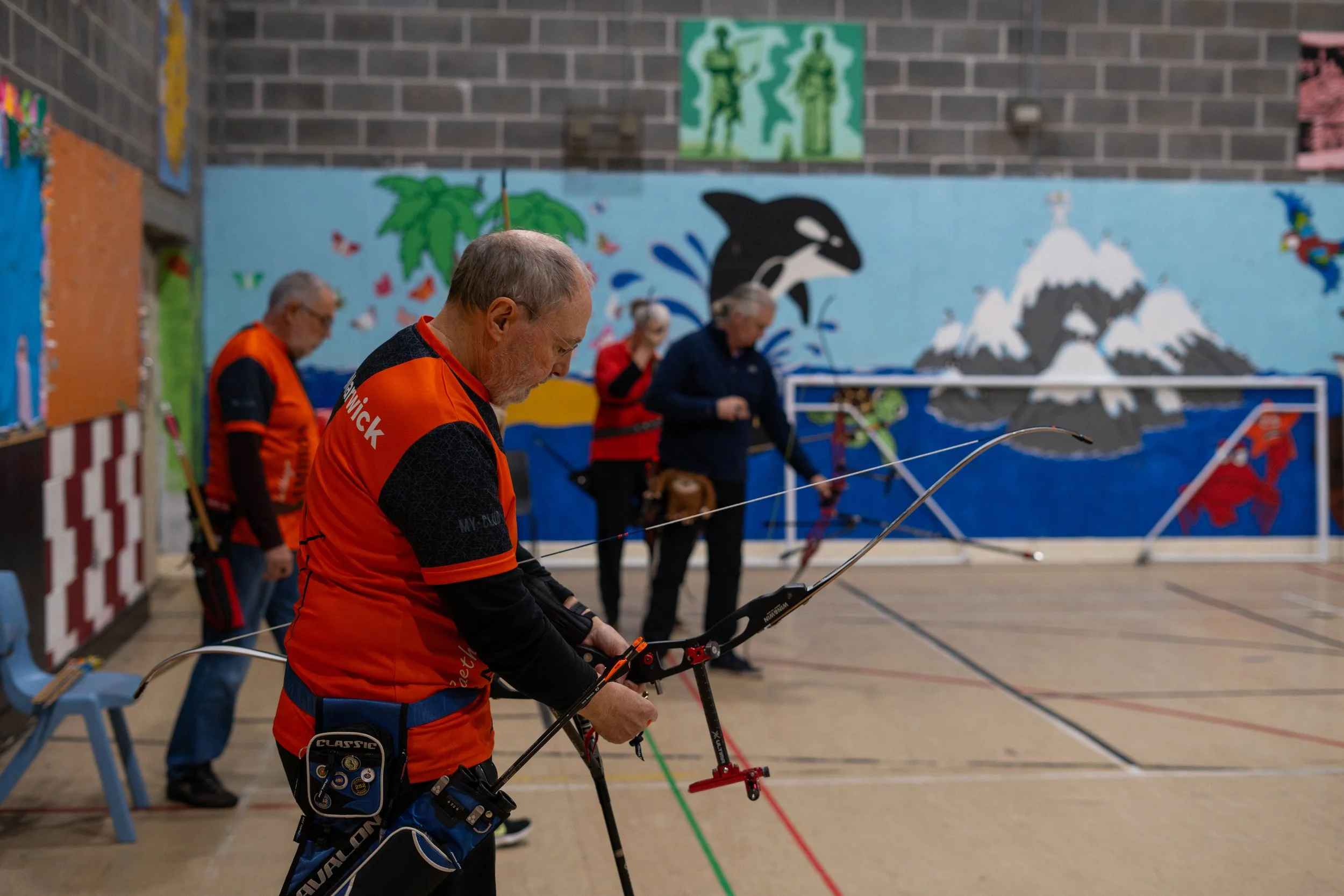 A group of senior people practicing archery indoors with colorful wall murals of mountain scenery, a whale, and a red dragon in the background.