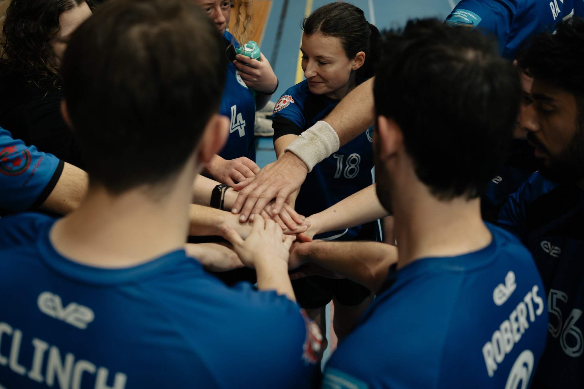 A team of volleyball players in blue jerseys huddles together with their hands stacked in the center, likely during a team meeting or pep talk.