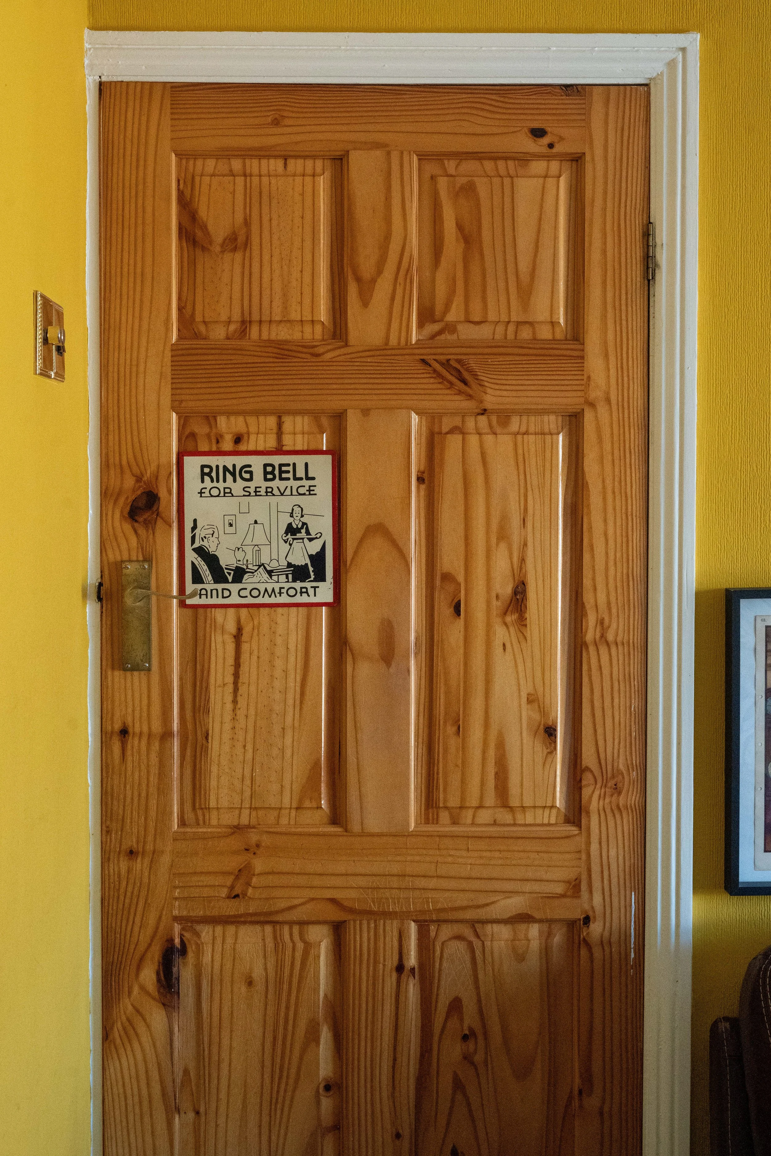 A wooden door with a sign hanging on it that reads, 'Ring Bell for Service and Comfort,' showing a person calling a bell and a person serving tea in a living room setting.