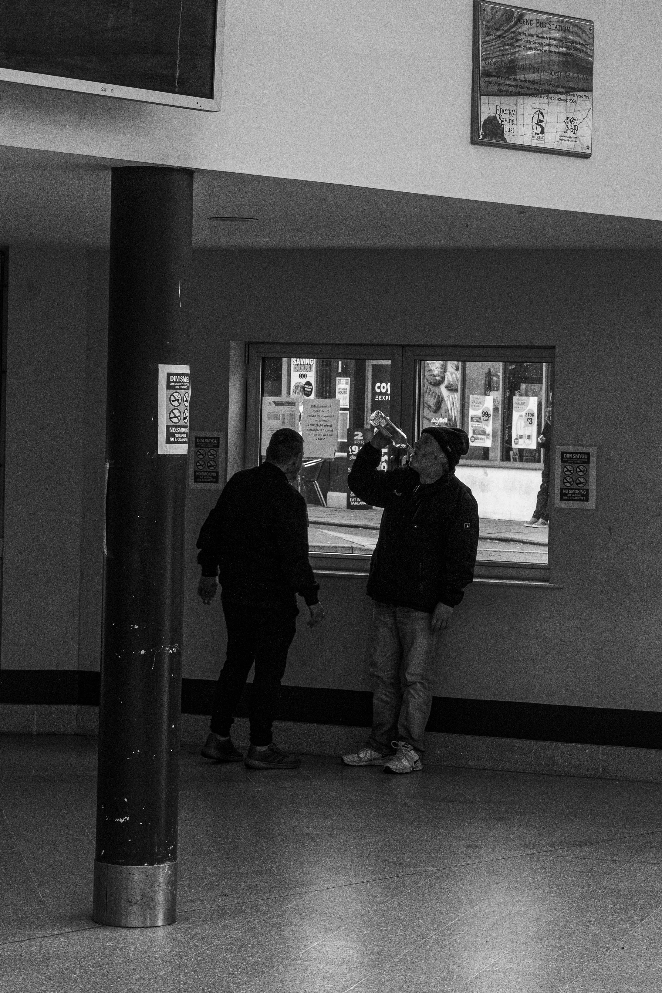 Two men inside a building, standing near a window. One man is drinking from a bottle, the other is facing away from the camera. The background shows a street scene with storefronts.