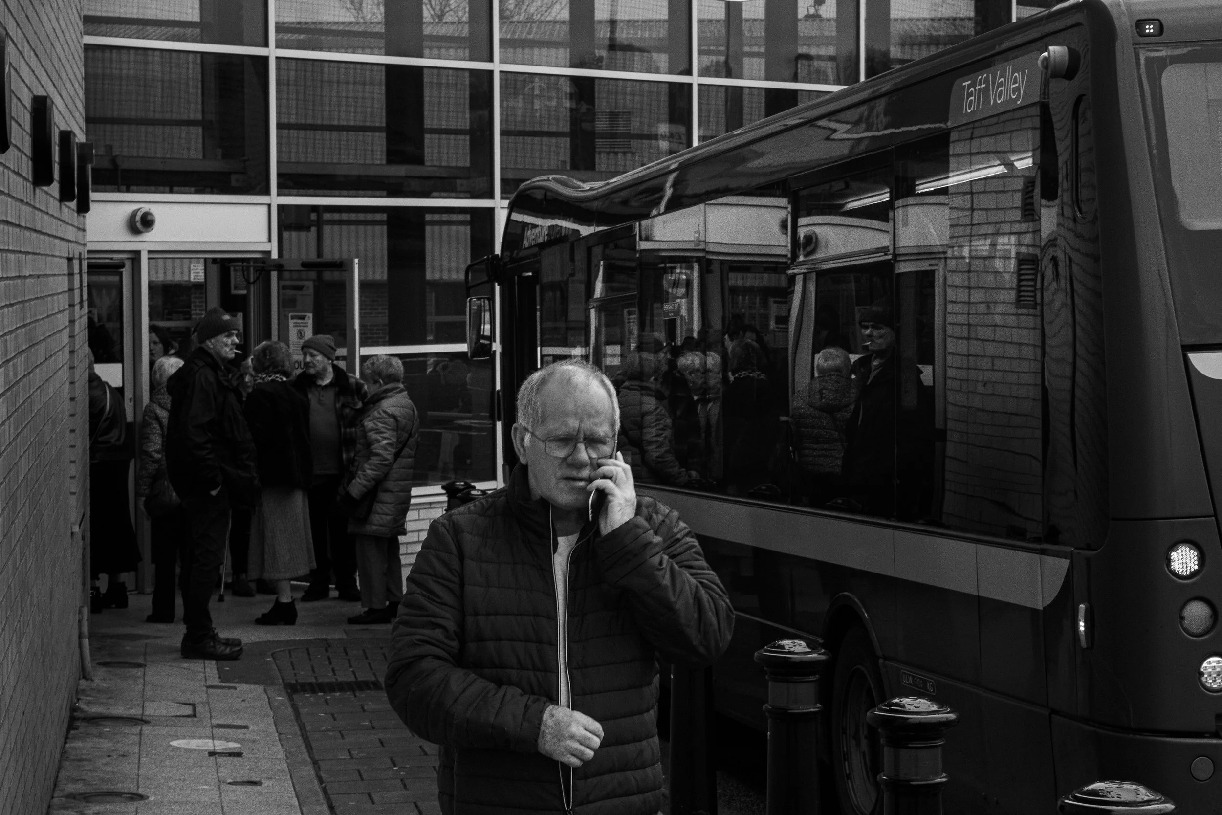 A man with glasses and a quilted jacket talking on a cellphone on a city sidewalk, with a bus labeled 'Taft Valley' and a group of people waiting outside a building behind him.