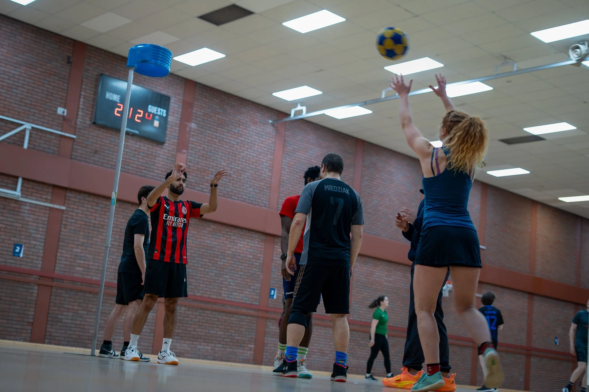 Group of people playing indoor volleyball in a gym, with a woman jumping to hit the ball over the net. The gym has brick walls and a digital scoreboard in the background.
