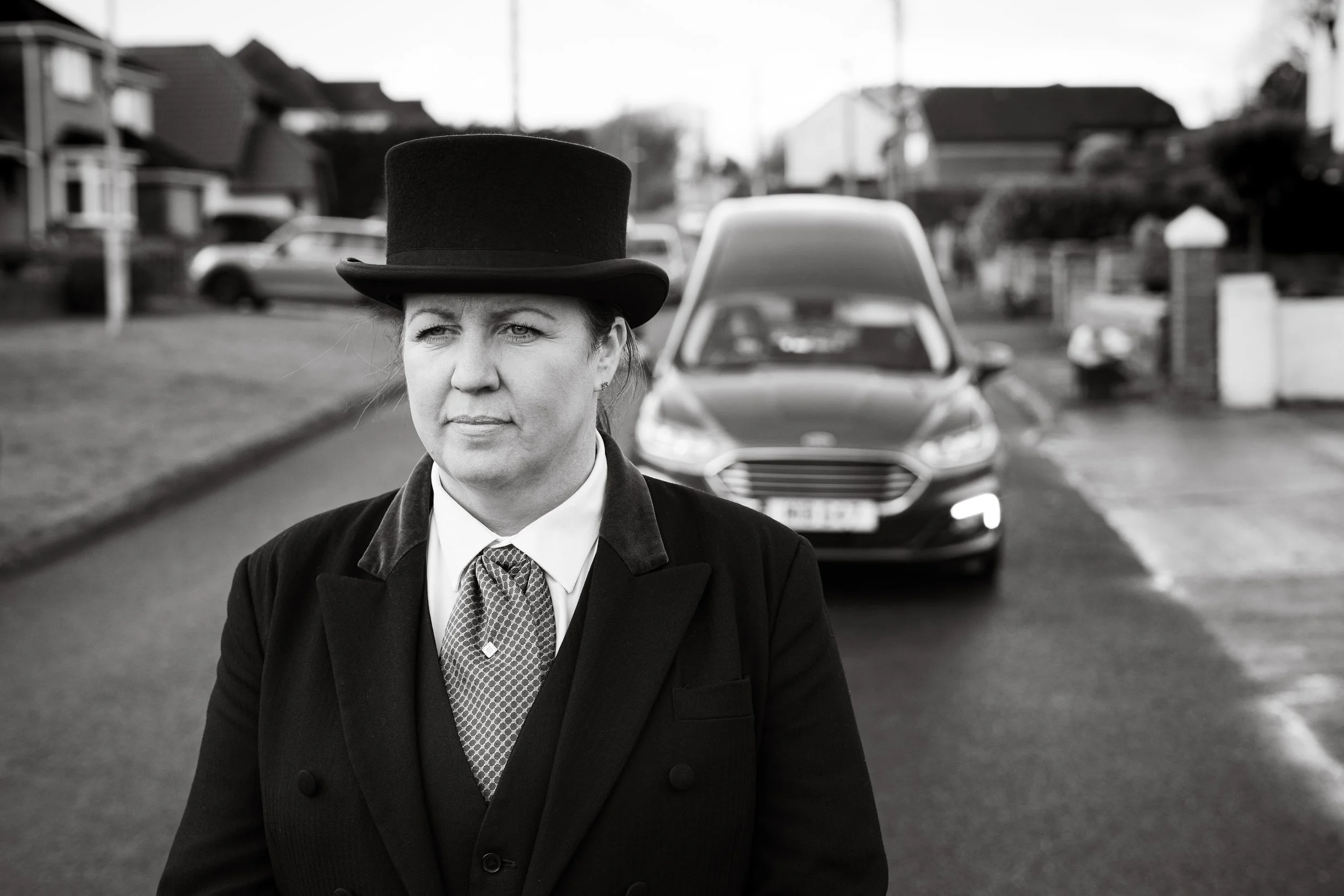 A woman dressed in vintage attire with a top hat and a coat, standing on a residential street with houses in the background, and a car parked behind her.
