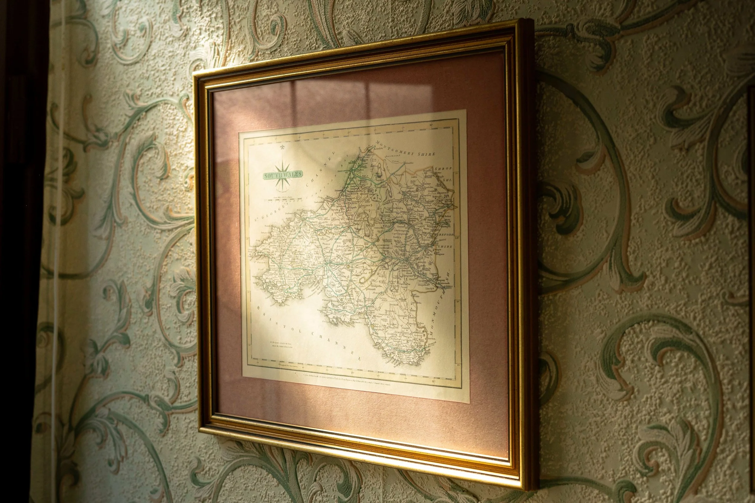 Framed vintage map of South Wales hanging on a decorative wallpapered wall.