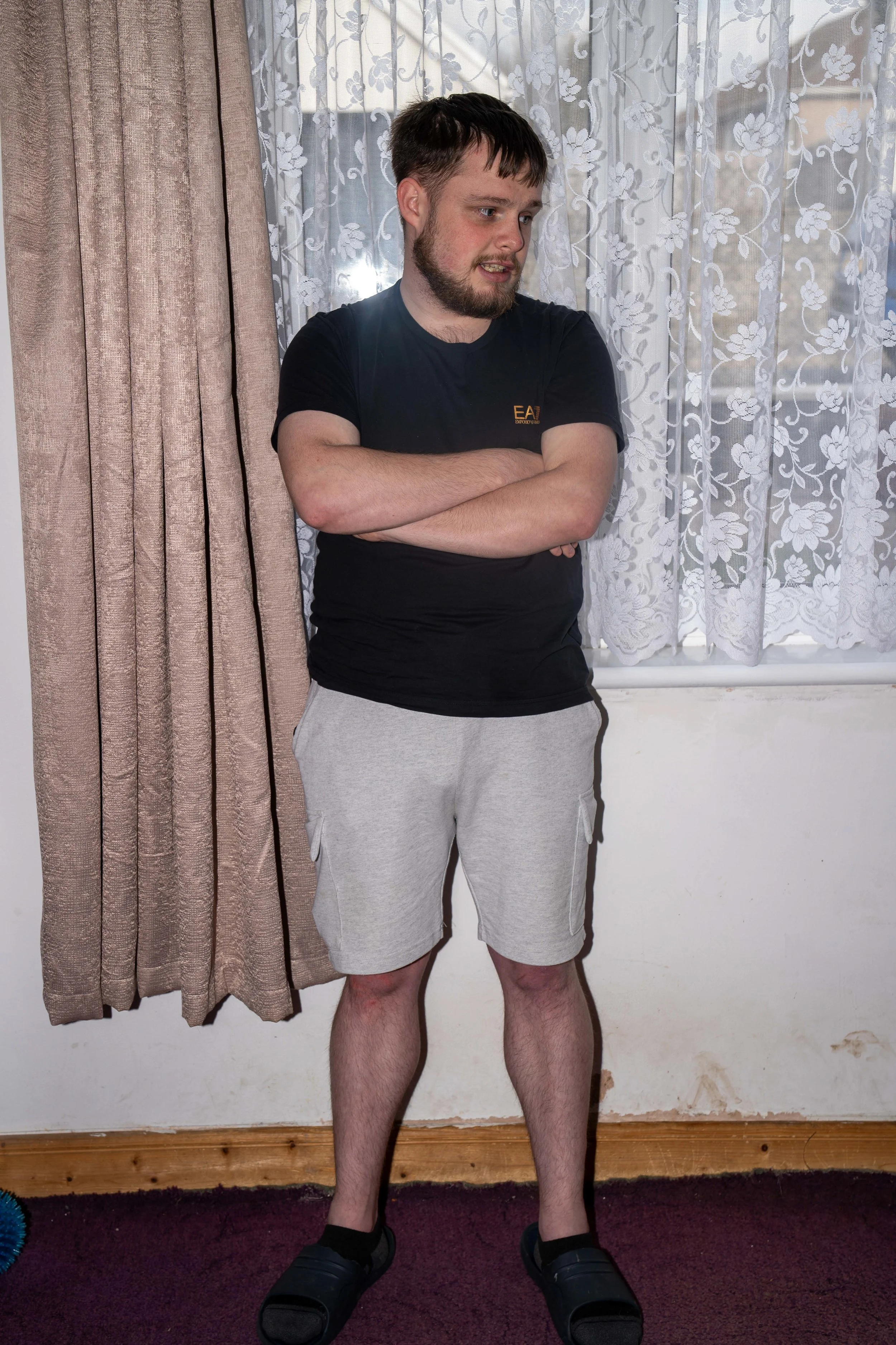 A young man with short dark hair and a beard, wearing a black t-shirt, gray shorts, and black slide sandals, standing with arms crossed and looking to the side inside a room. There are curtains and lace window treatments behind him.