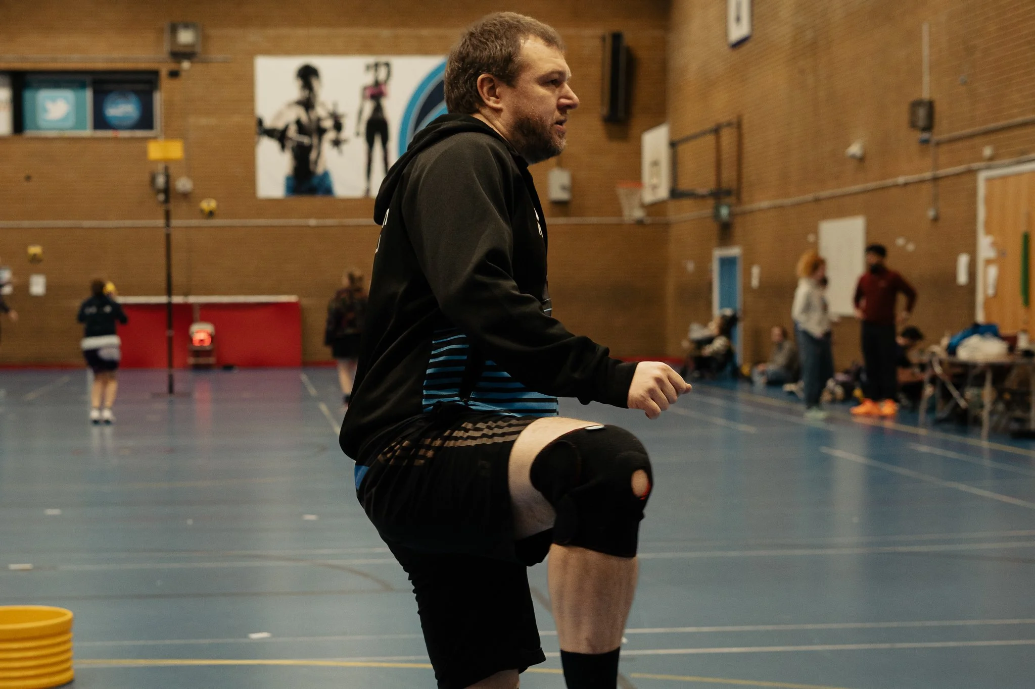 A man wearing a black jacket and black shorts with a knee brace is stretching in a gymnasium. The gym has a blue floor and a brick wall, with other people and sports equipment in the background.