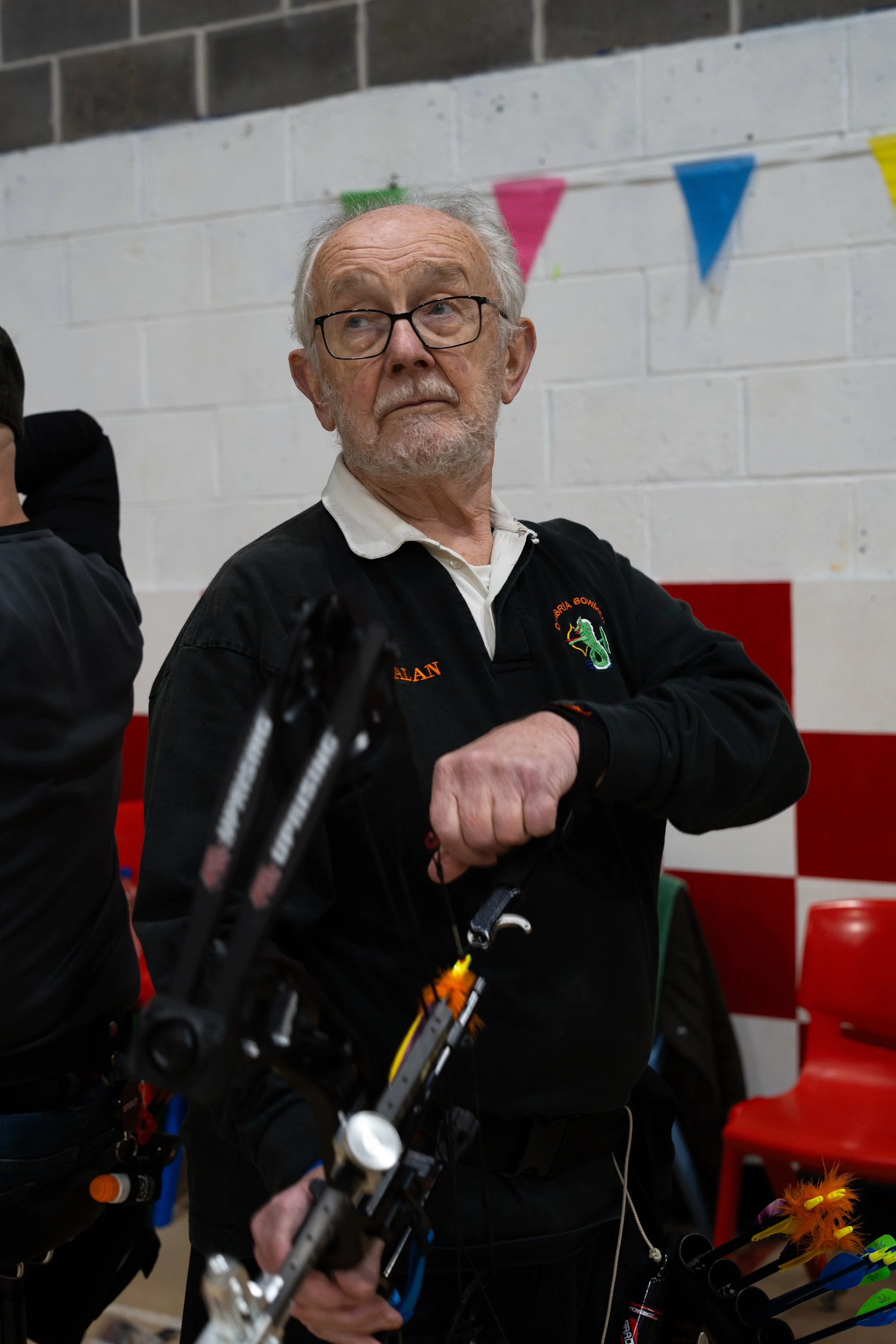 An elderly man with glasses and a white beard wearing a black jacket with a store logo and the name 'ALAN' patch, standing with crossed arms at an indoor archery event, holding a bow, with colorful bows and arrows visible in the foreground.