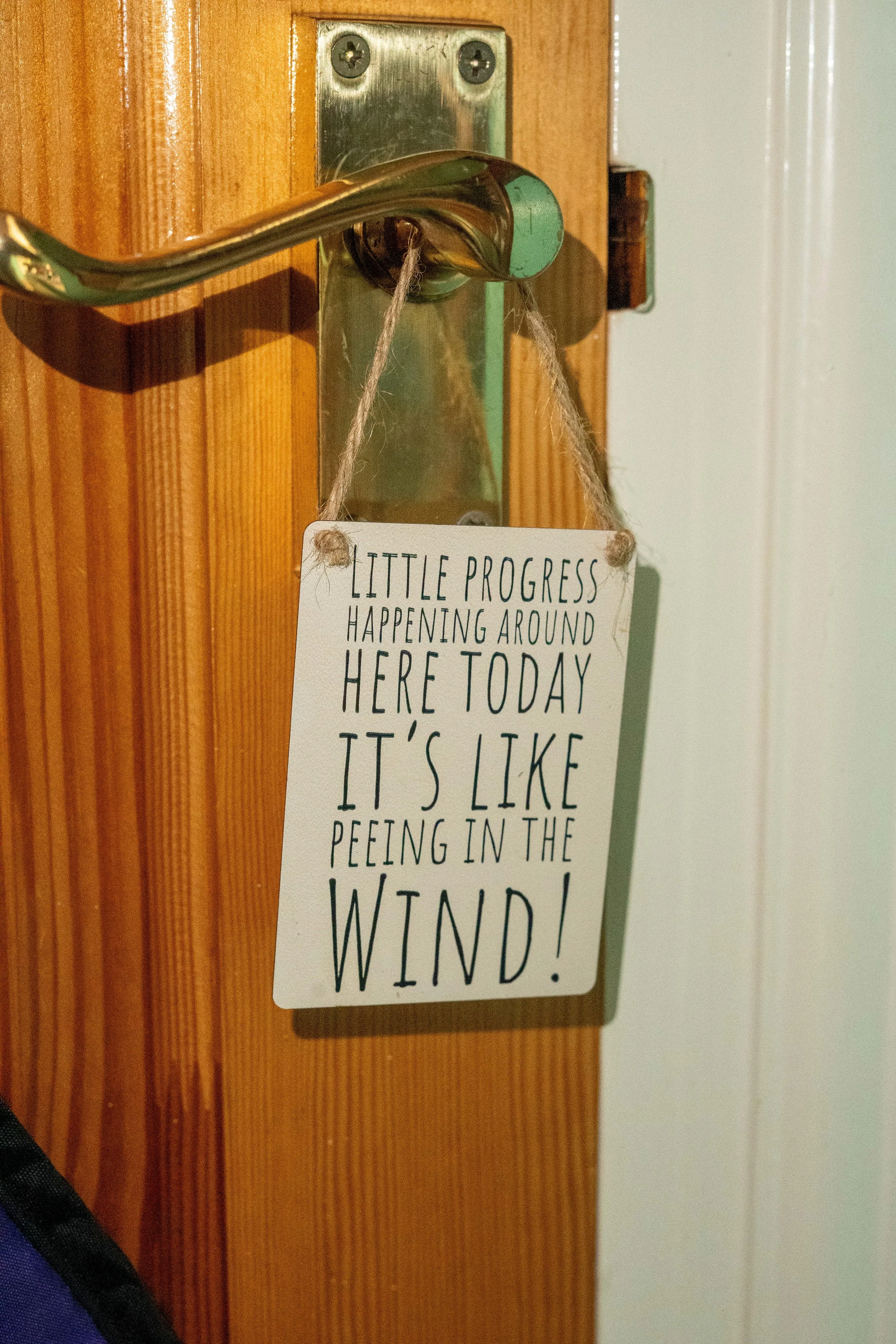 A wooden door with a shiny brass door handle and an attached sign that reads "Little progress happening around here today, it's like peeing in the wind!"