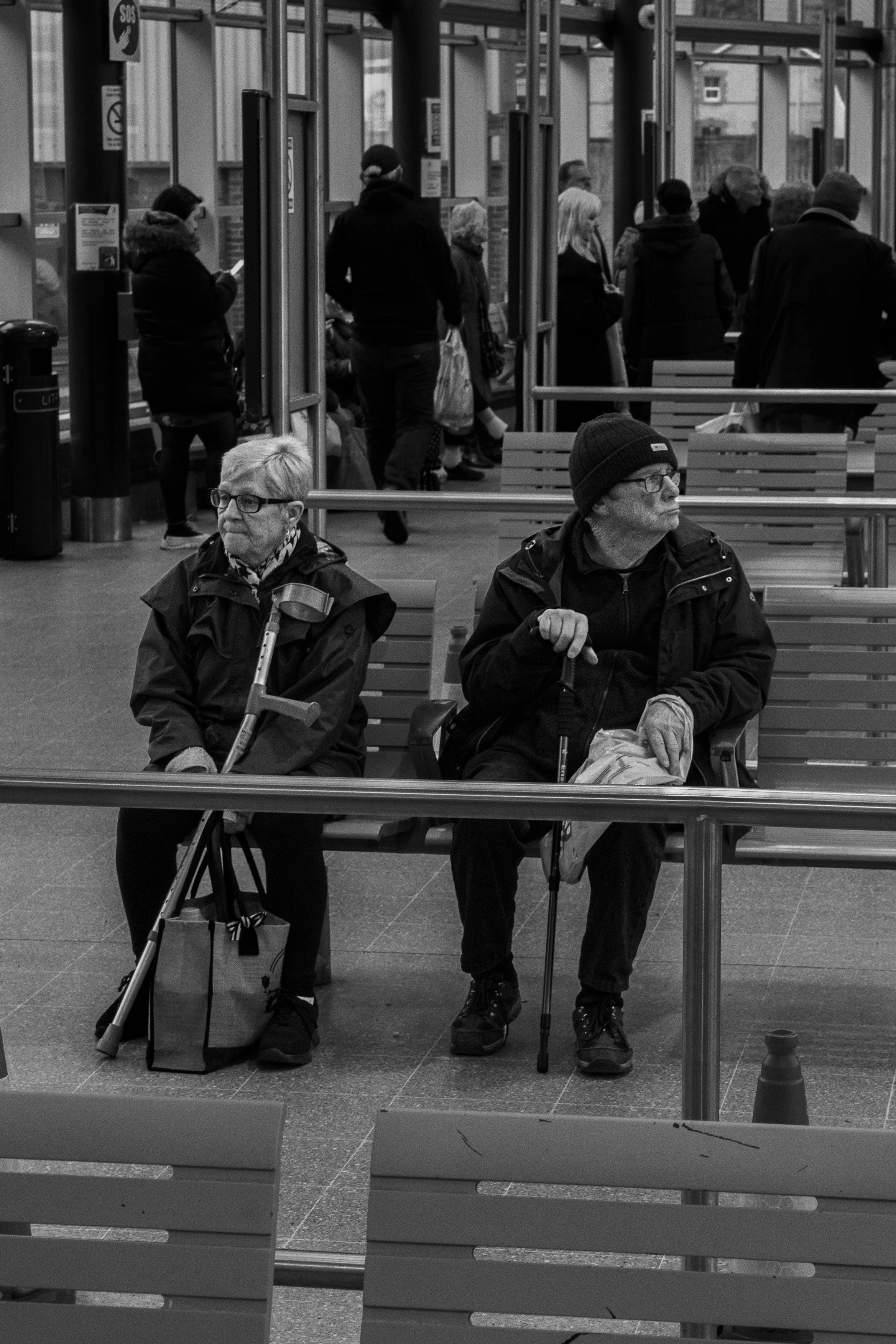 Two elderly individuals sitting on a bench at a public transportation station, with a group of people standing and walking behind them in black and white.