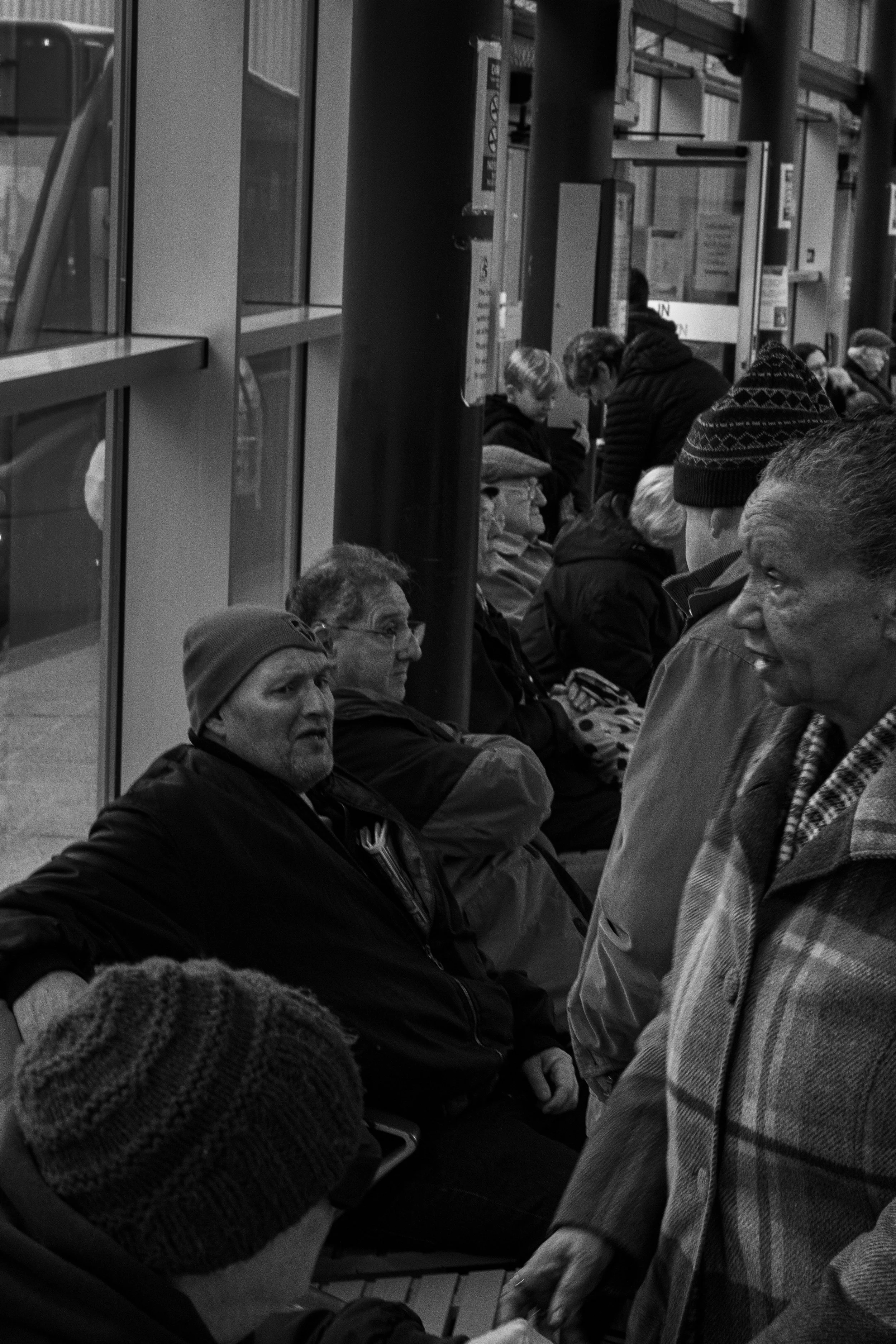 People sitting and waiting on benches at a transit station or bus stop, some engaged in conversation.