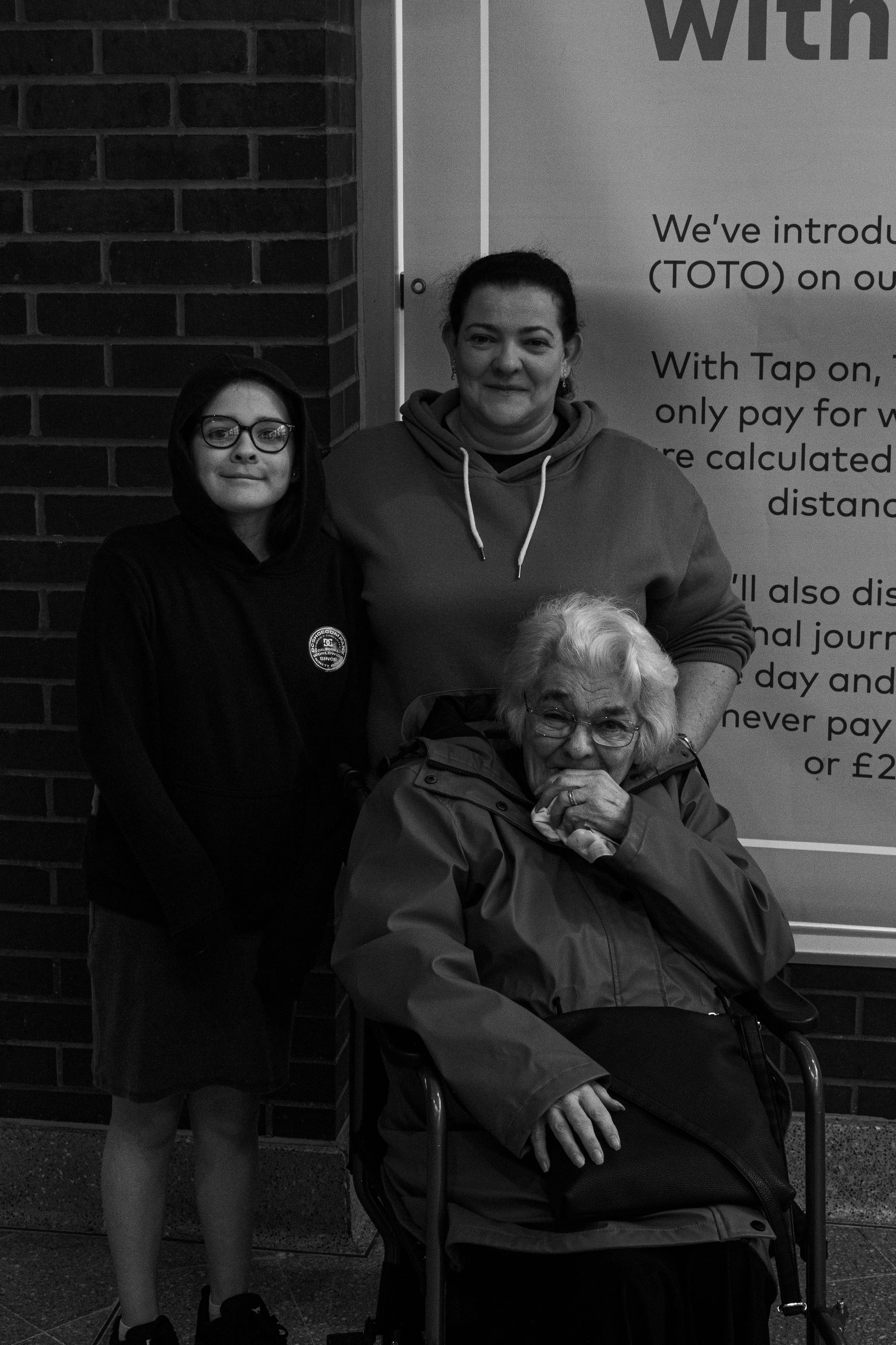 Three women standing together, one elderly woman sitting in a wheelchair, outside a building with a sign in the background.