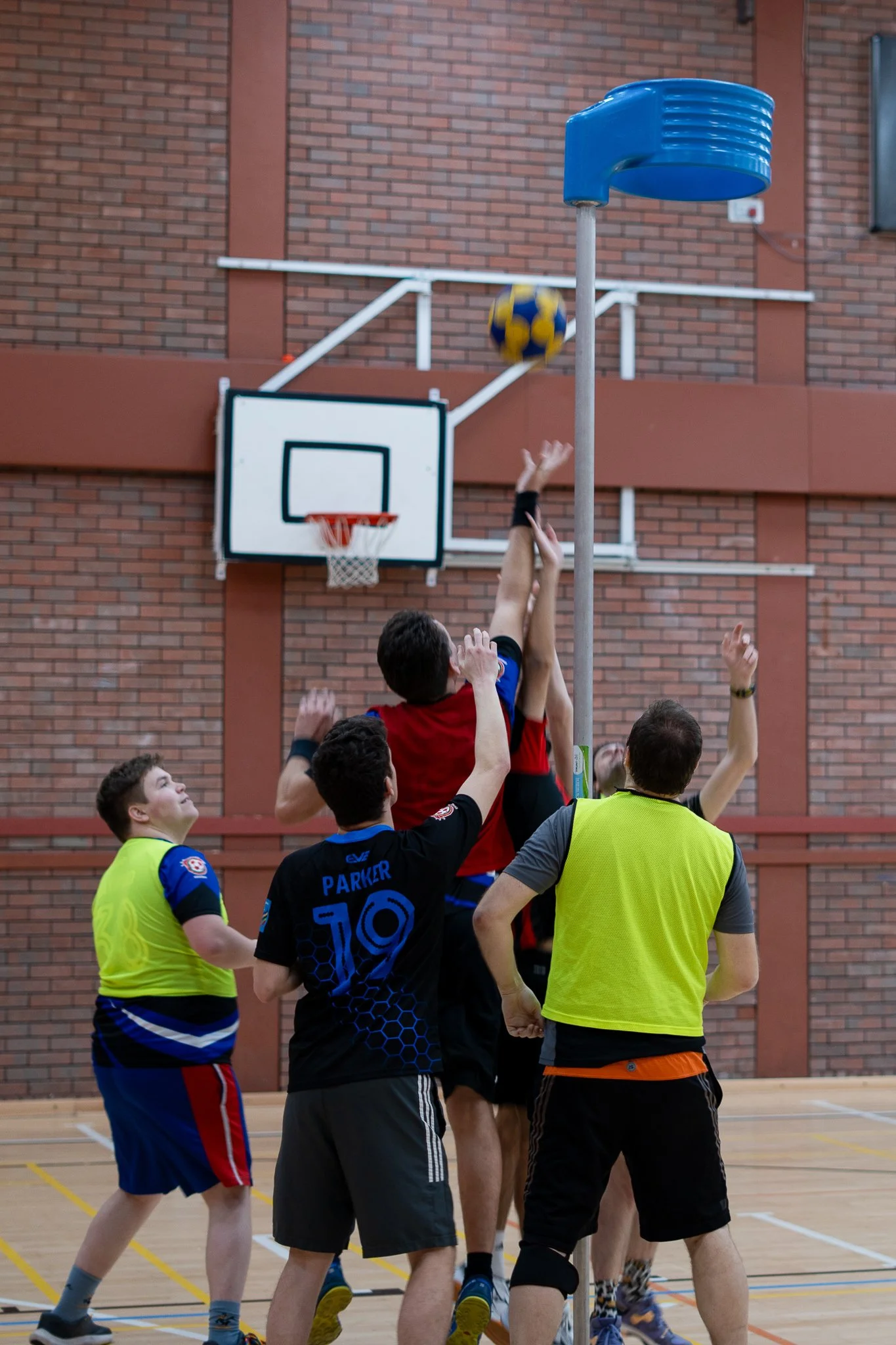 Group of men playing basketball indoors, jumping for ball near hoop and basketball pole.