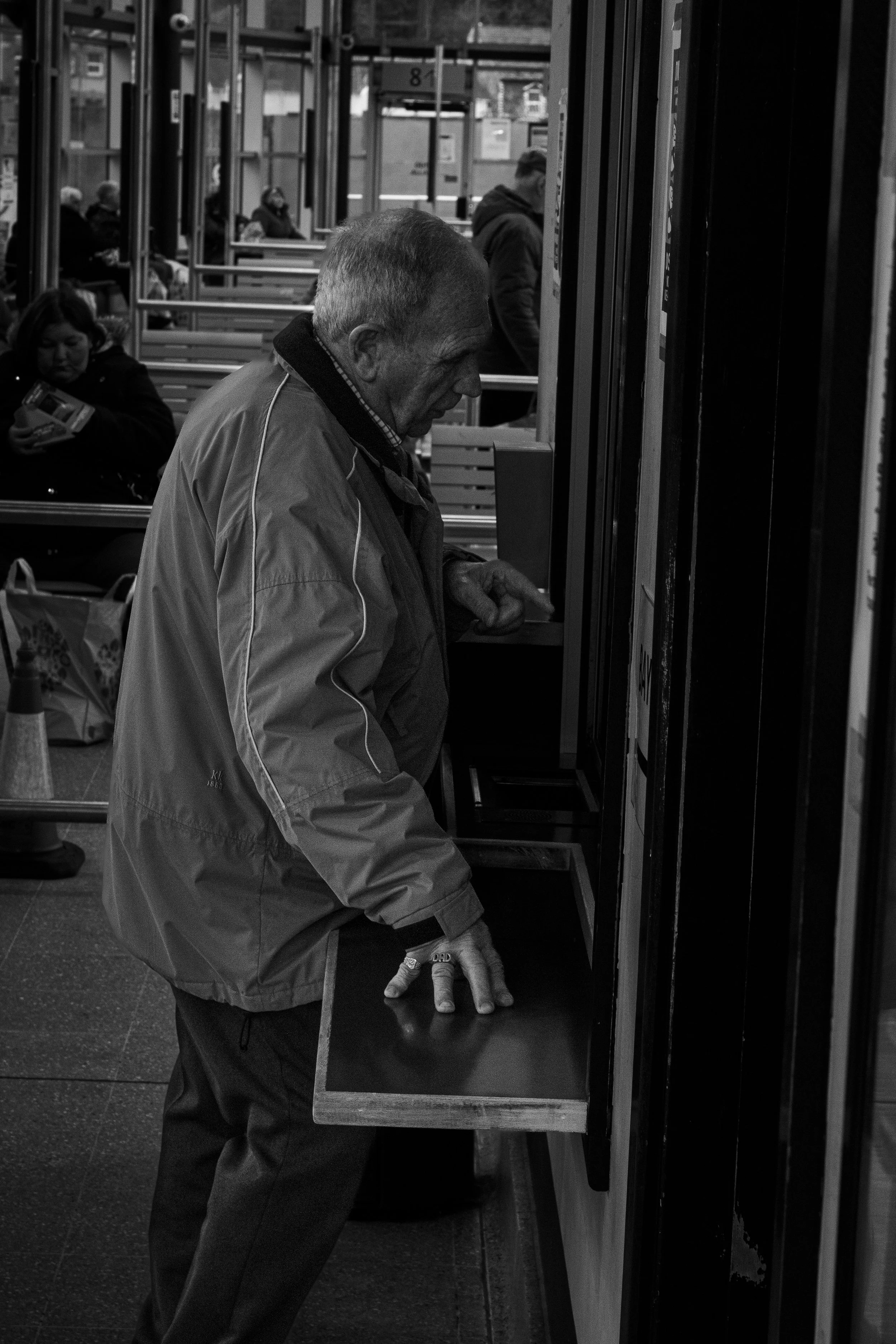An elderly man in a jacket is at a ticket machine, pressing buttons. People are seated in the background at a transportation station.