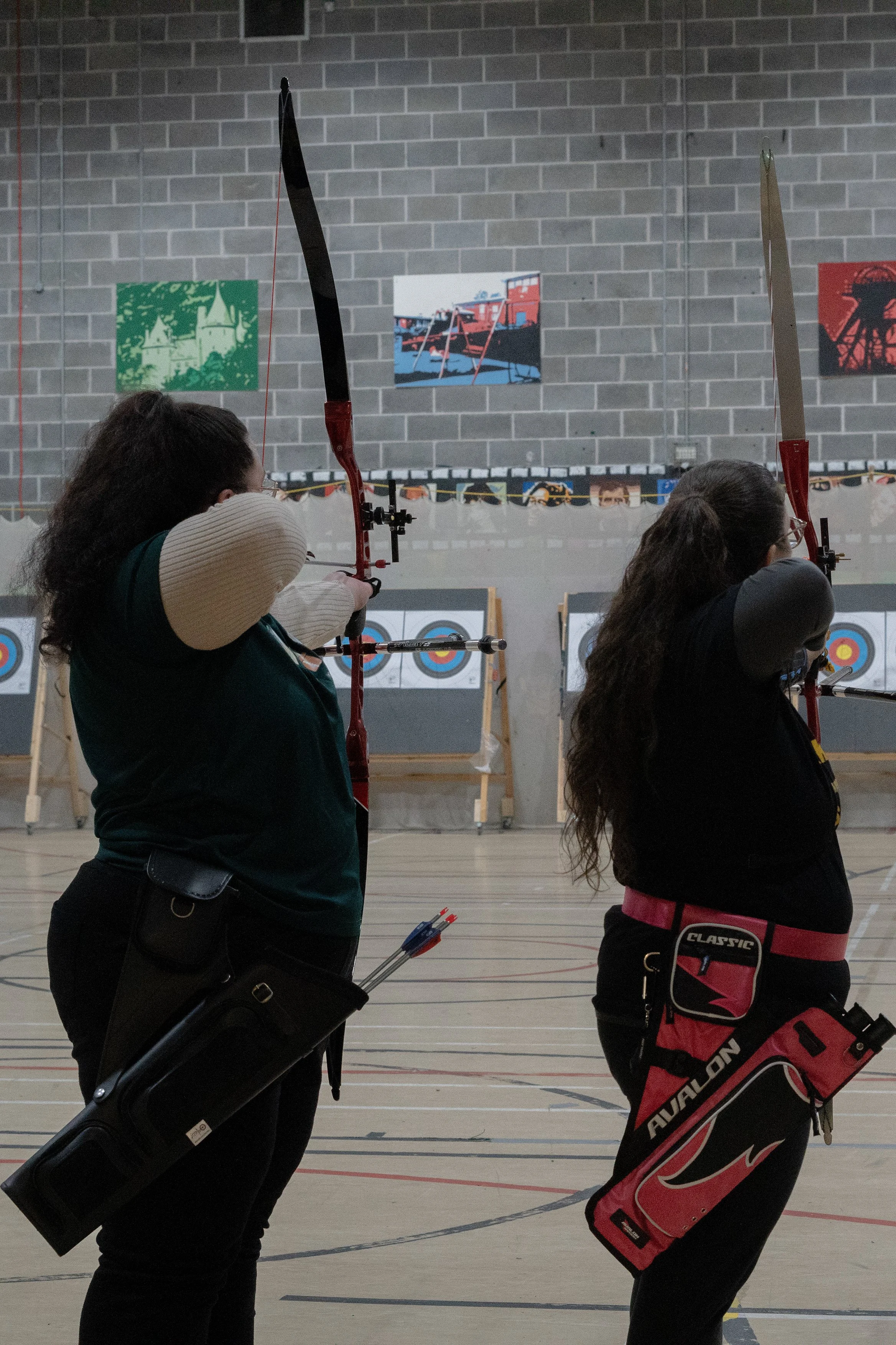 Two women practicing archery indoors, aiming with bows and arrows, with archery target boards in the background.