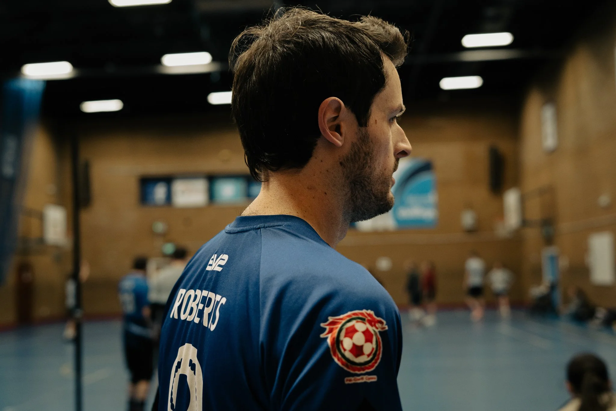 A man in a blue sports jersey with the name 'Roberts' on the back, standing in an indoor sports hall with other players in the background.