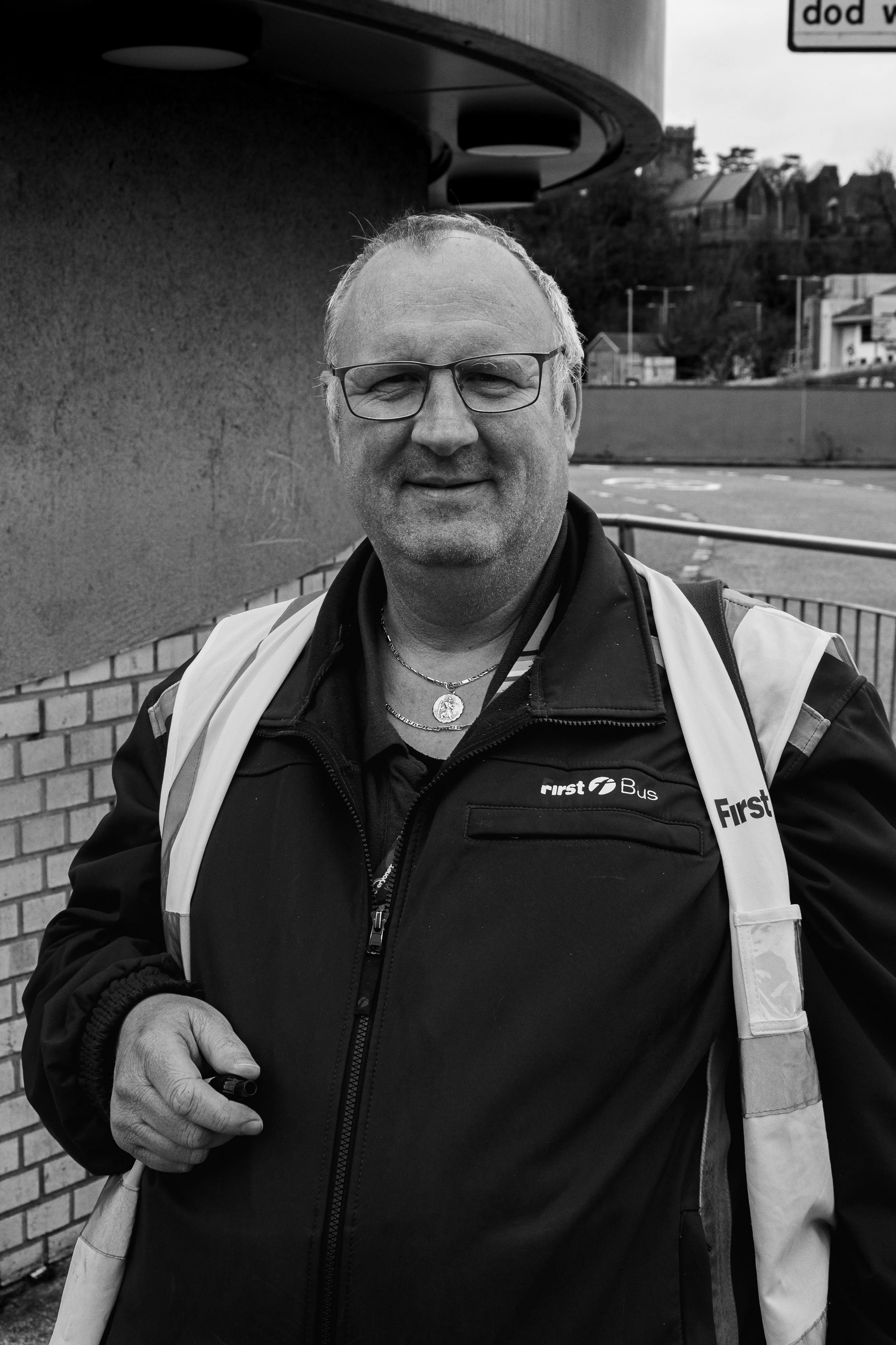 A man wearing glasses and a First Bus uniform, standing outdoors near a brick wall and a bus stop.