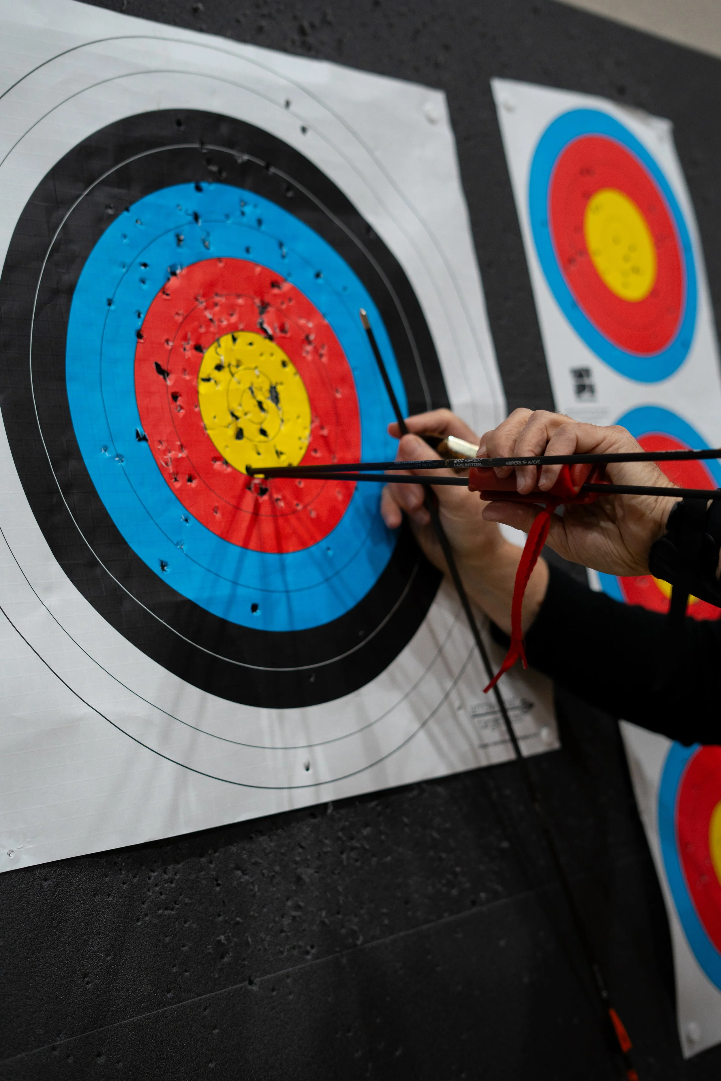 A person is holding an arrow and pointing it at the bullseye of a target with multiple bullet holes, with other similar targets in the background.