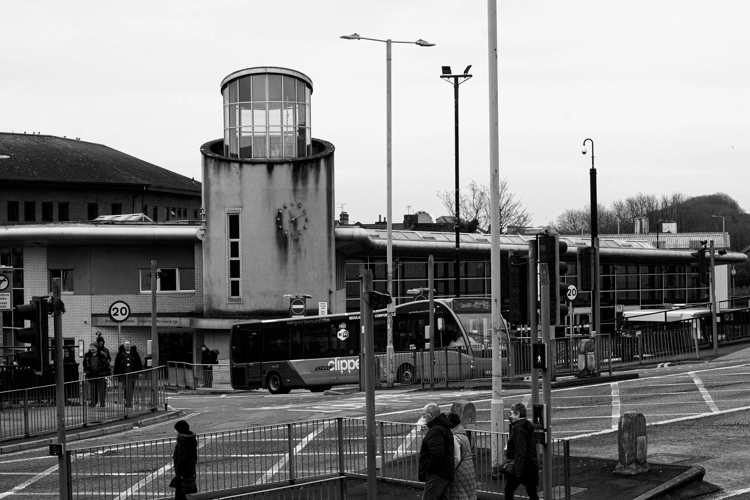 A black and white photo of a bus stop with people walking and waiting, a bus parked at the station, and a distinctive cylindrical building with a clock near the top.