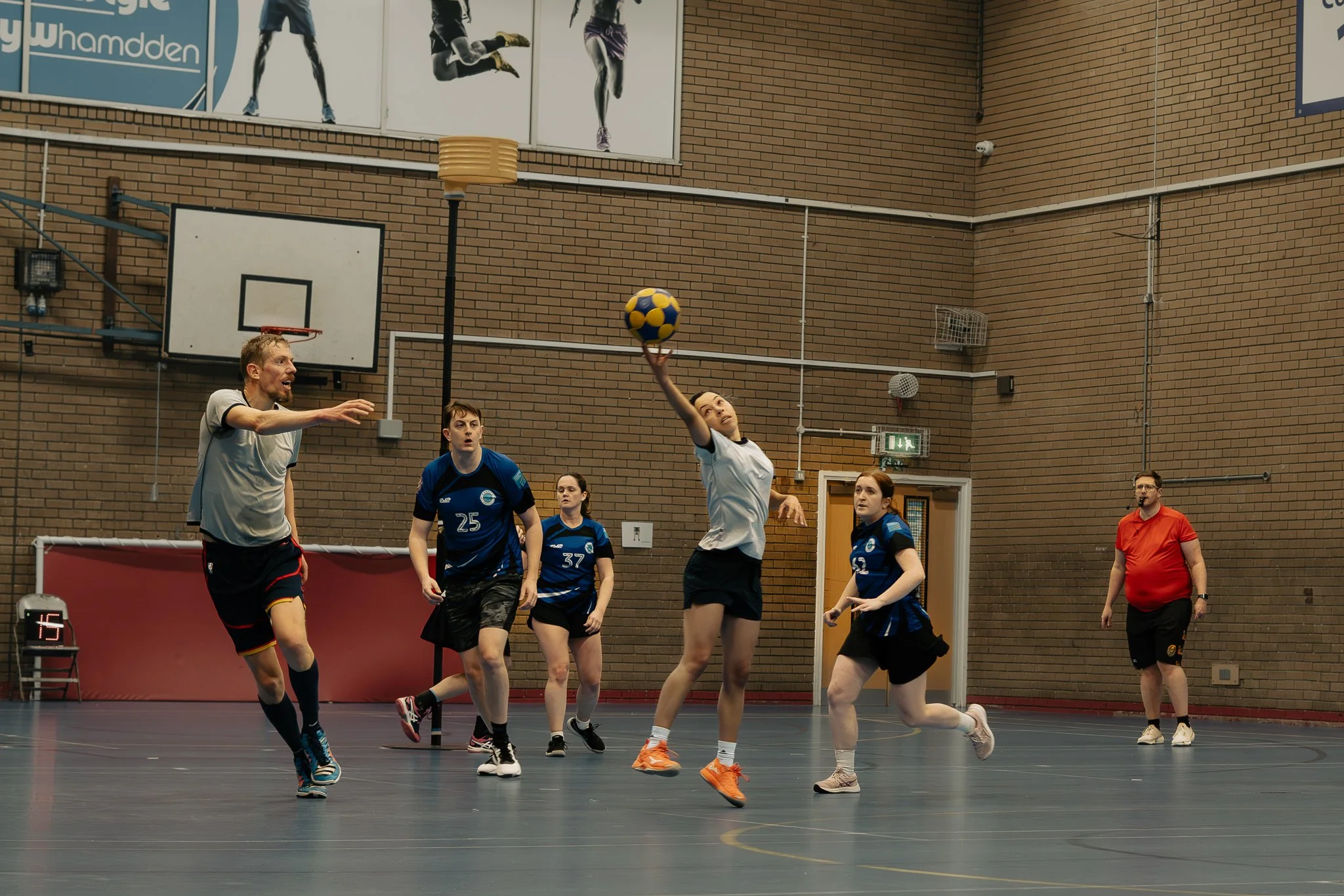 A group of people playing indoor handball on a court. One woman is jumping to throw a ball, while others are preparing to block or catch. A man stands on the sideline watching.