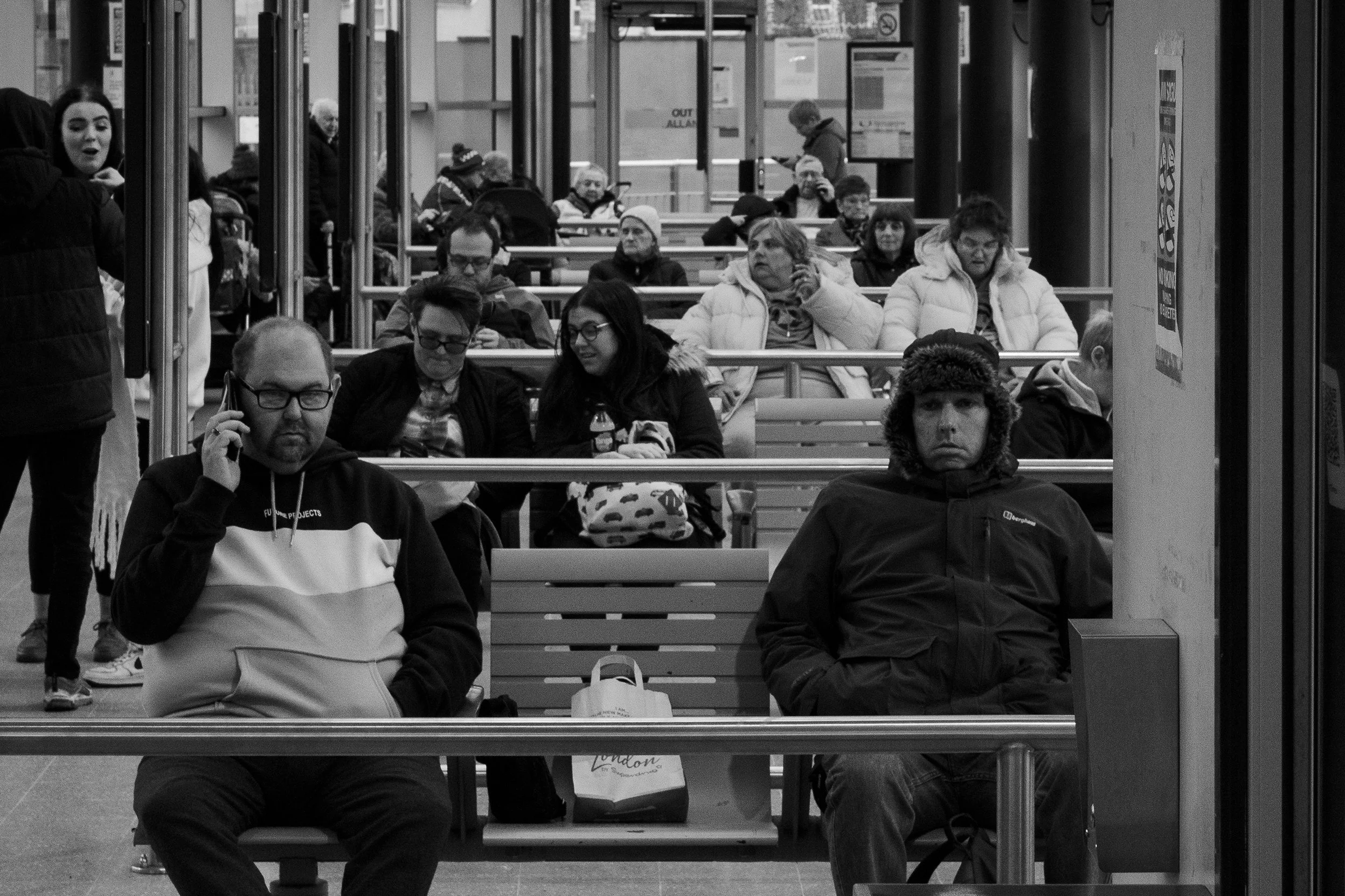 People sitting on benches at a busy transportation hub, with some using phones and others waiting.