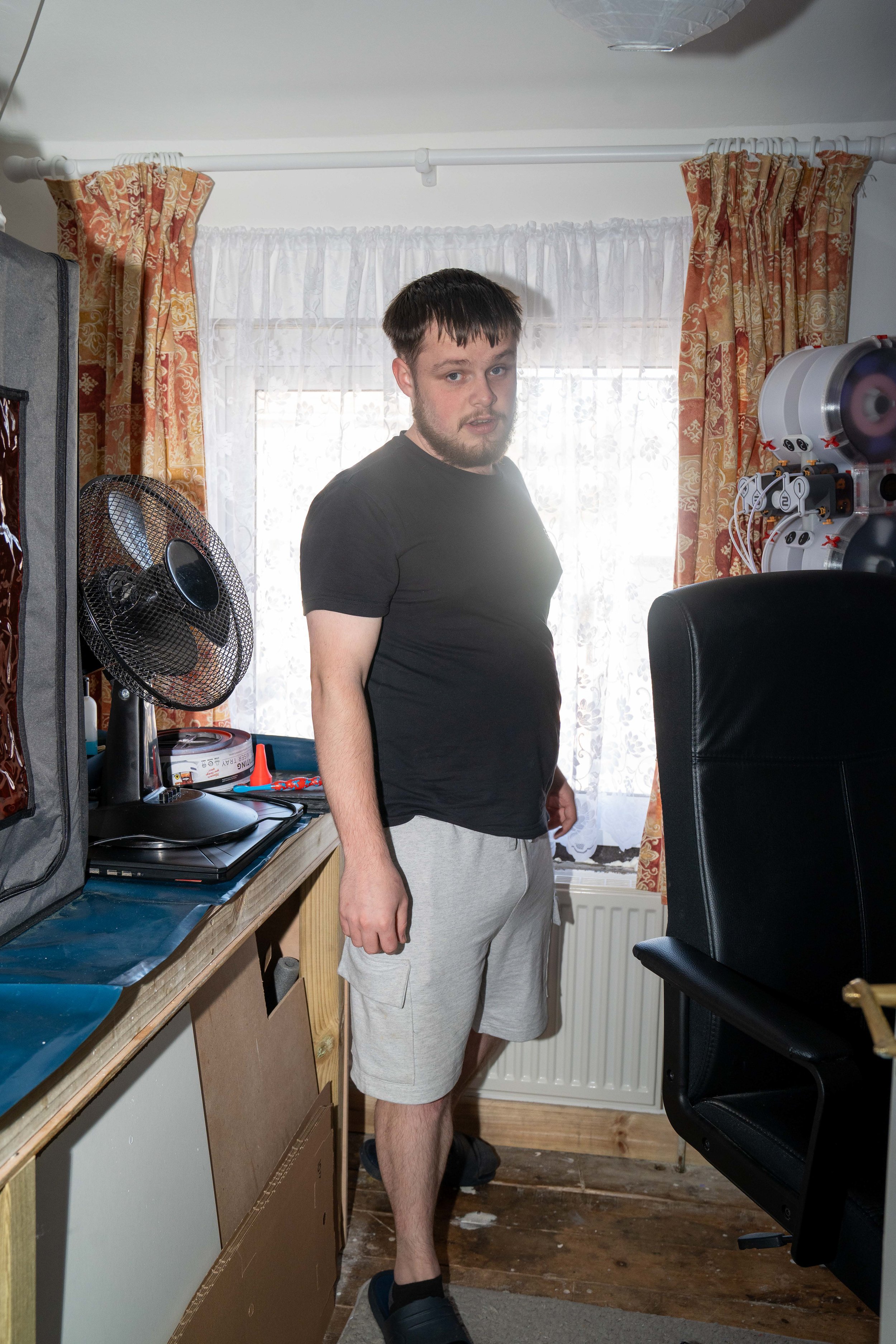 A young man with dark hair and a beard standing in a room near a desk with a fan and other items, in front of a window with sheer curtains and floral drapes, wearing a black t-shirt and light gray shorts.