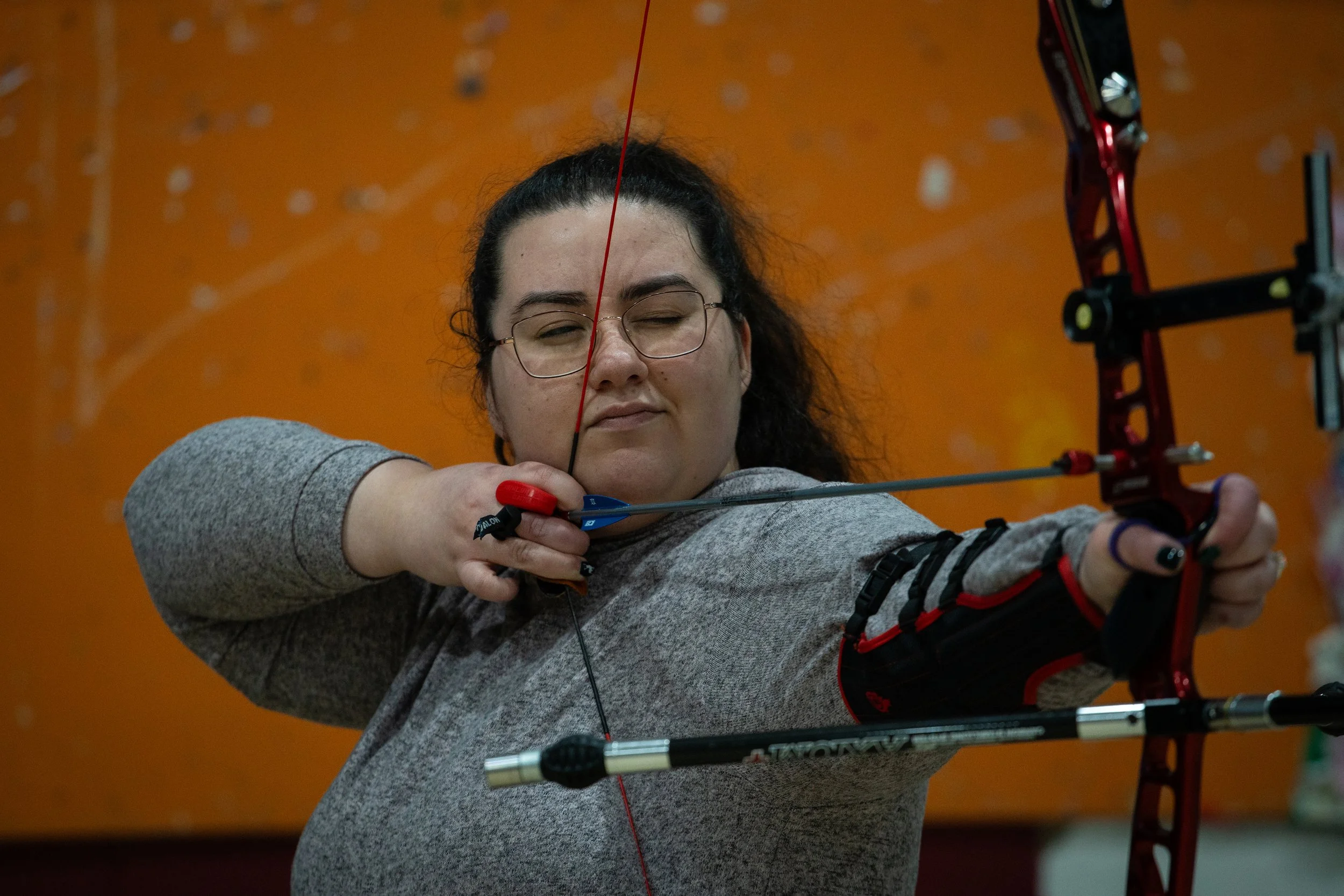A woman with glasses practicing archery indoors, aiming with a red and black bow against an orange wall background.