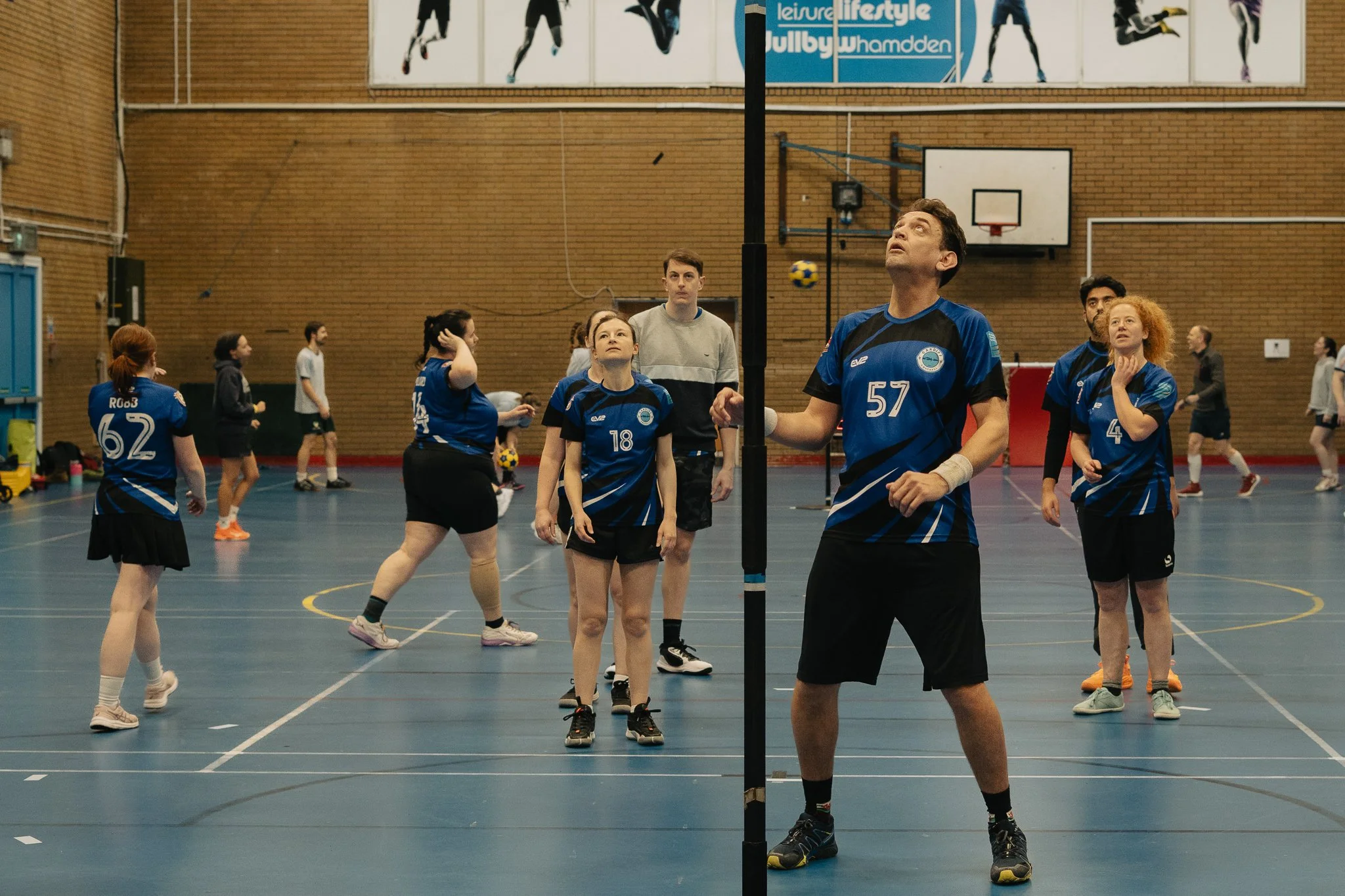 An indoor volleyball game with players on both sides of the net, some preparing to serve or receive, in a gymnasium with a brick wall and a basketball hoop in the background.