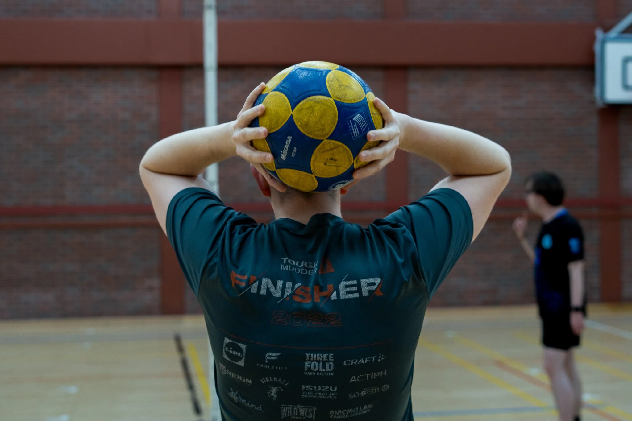 Person holding a yellow and blue water polo ball behind their head on an indoor sports court, with another person standing in the background.