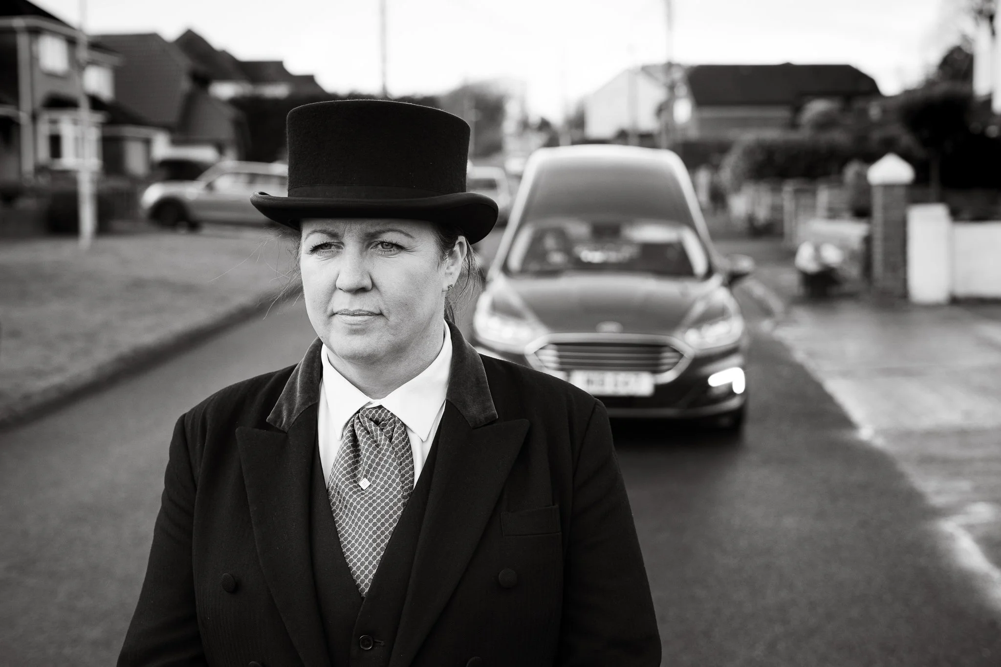 A woman dressed as a Victorian-era character, wearing a top hat and coat, standing on a suburban street with houses and a modern car in the background.