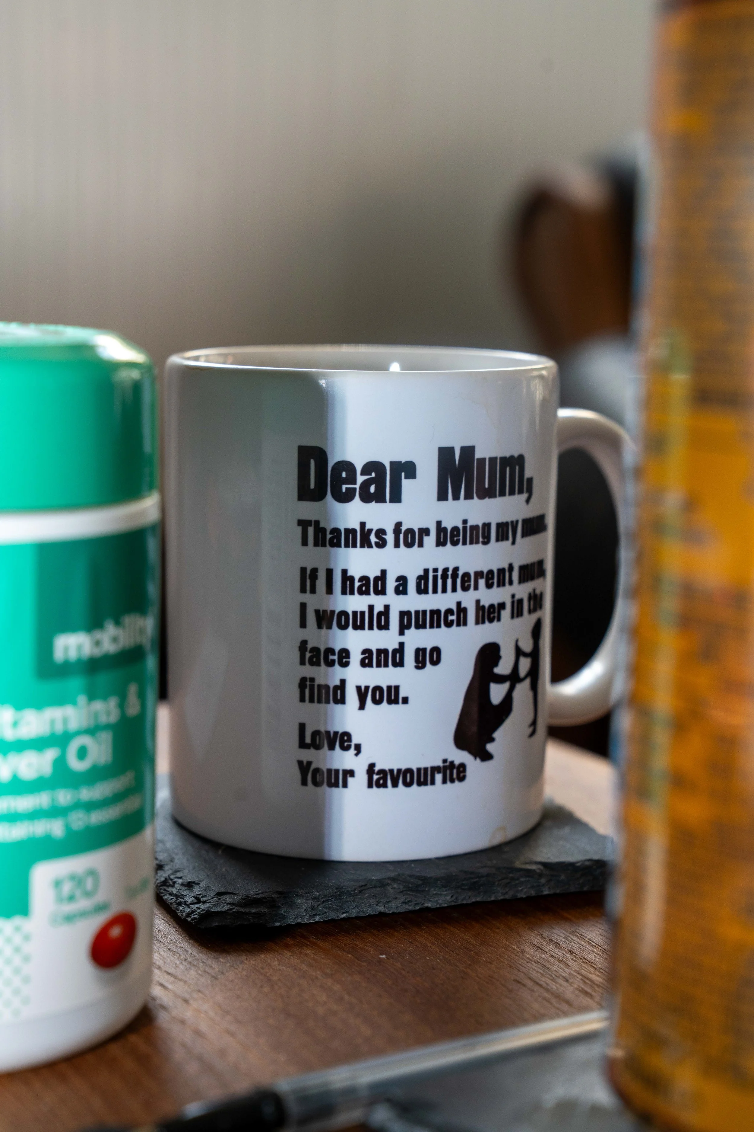 A coffee mug with a humorous message about mothers, placed on a wooden table surrounded by other containers and objects.