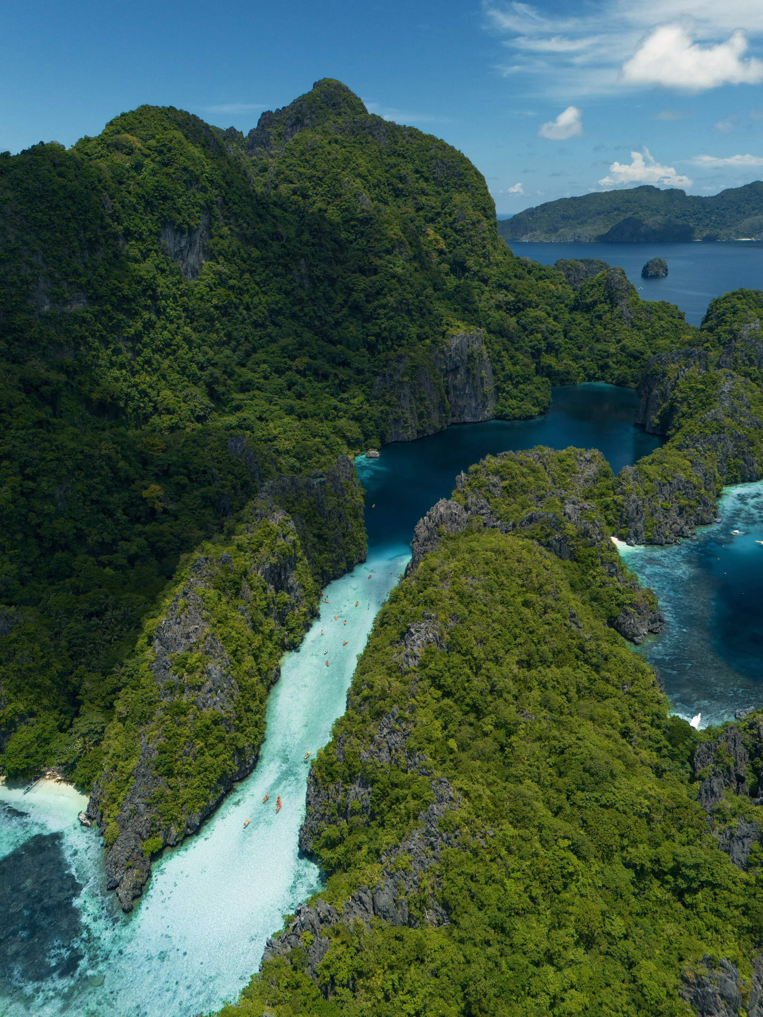 Aerial view of a tropical landscape with lush green islands, turquoise waters, and small boats nearby, under a blue sky with some clouds.