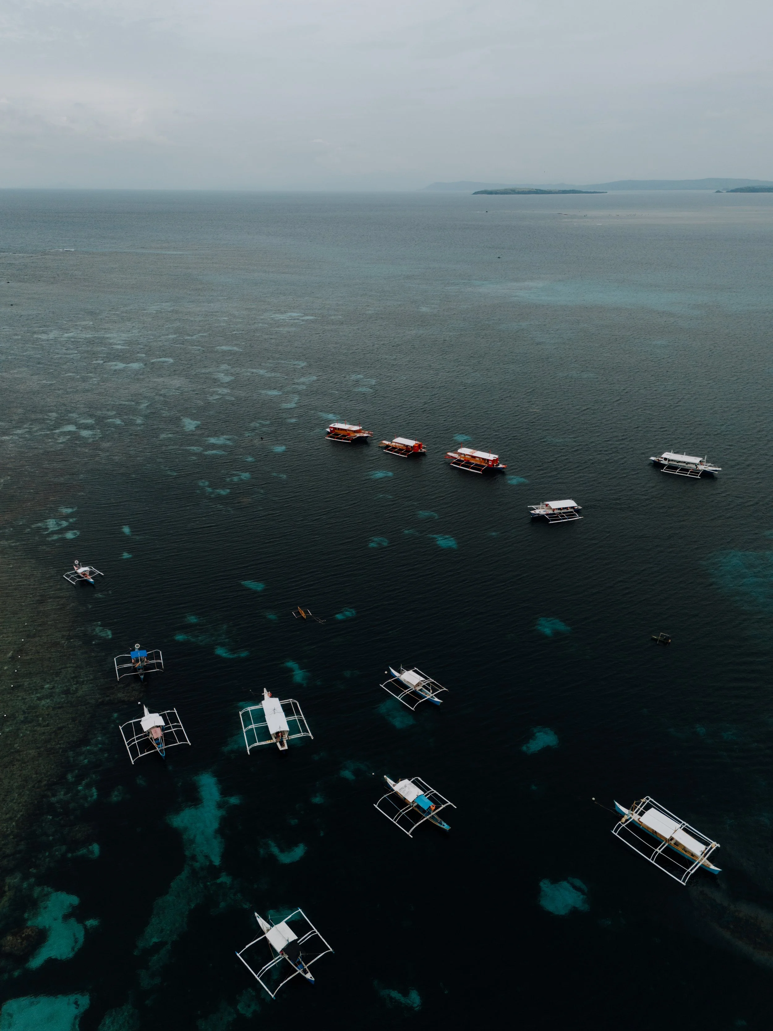Aerial view of several traditional boats floating on dark ocean waters with visible coral reefs underneath, under cloudy sky.