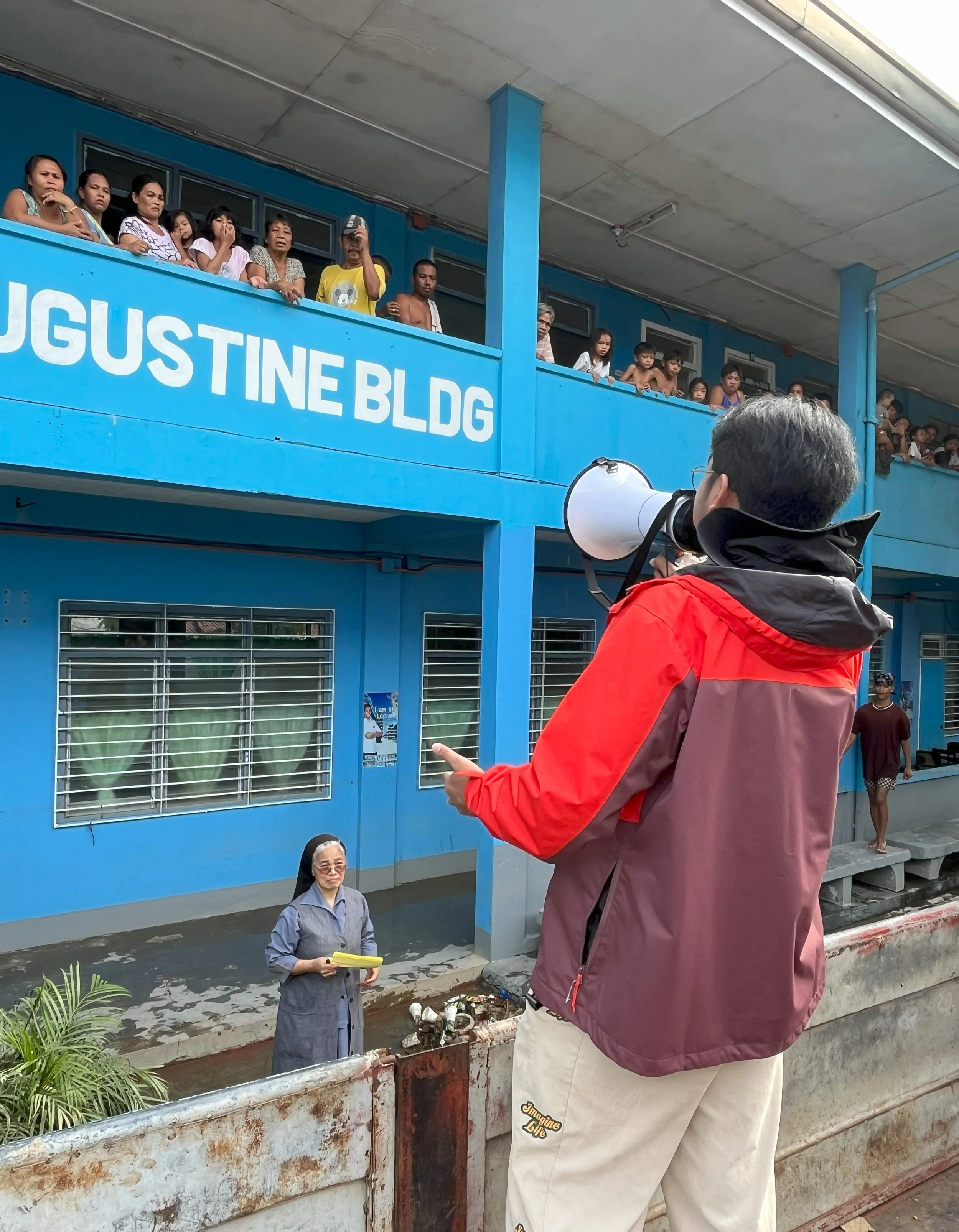 A man in a red and gray jacket speaking into a megaphone, standing in front of a blue building labeled "Augustine BLDG," with people on the second floor observing, and a woman in a gray dress and glasses standing outdoors at the building's entrance.