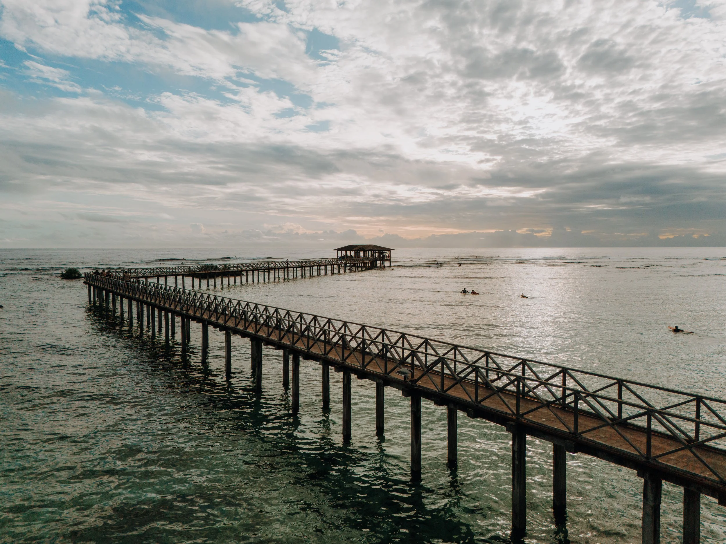 A long wooden pier extends over the ocean, leading to a small covered structure. Several people are on the pier and in the water, with a cloudy sky overhead.
