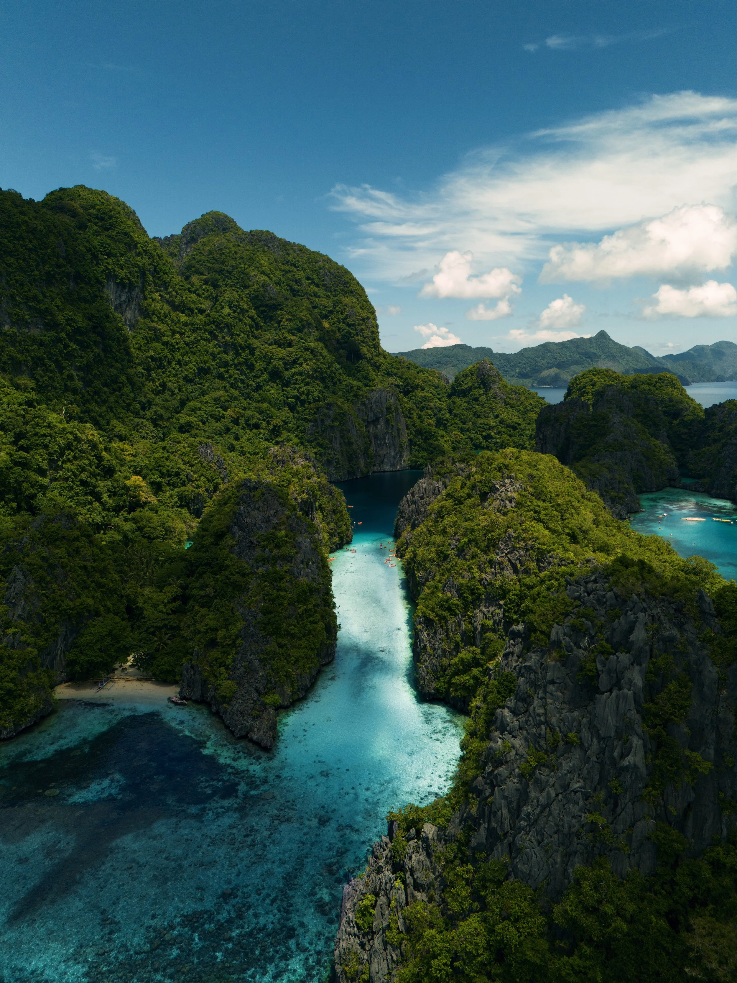A landscape of lush green mountains surrounding a narrow turquoise waterway with small boats, under a blue sky with some clouds.