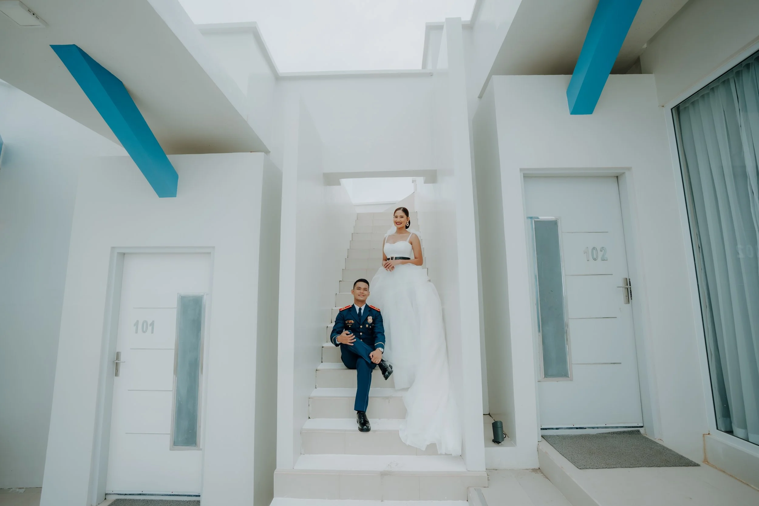 A man in a blue military uniform and a woman in a white wedding dress sitting on white stairs inside a modern white building with blue accents.