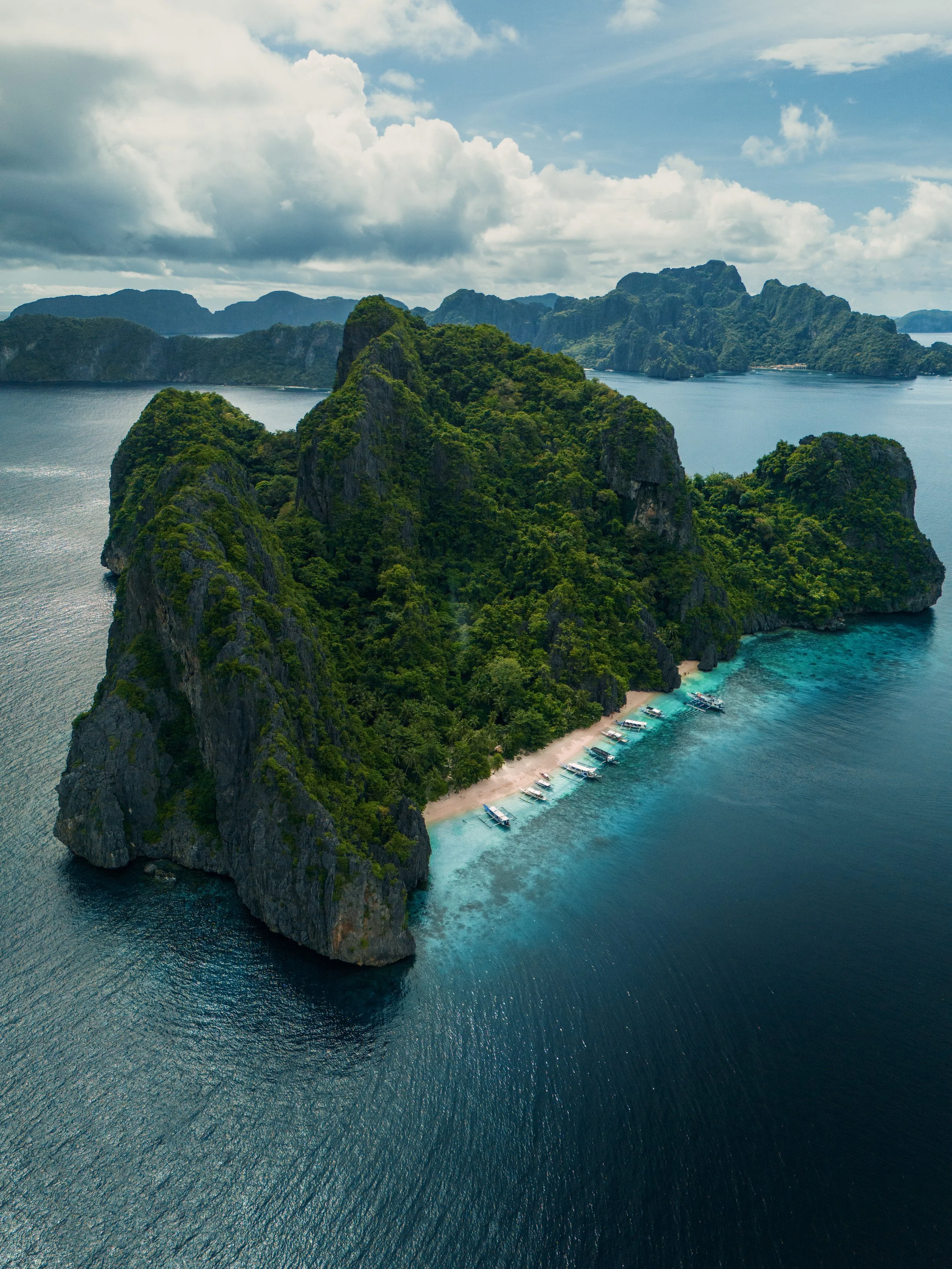 Aerial view of a lush, green island surrounded by turquoise water, with boats docked near a sandy beach, under a partly cloudy sky.