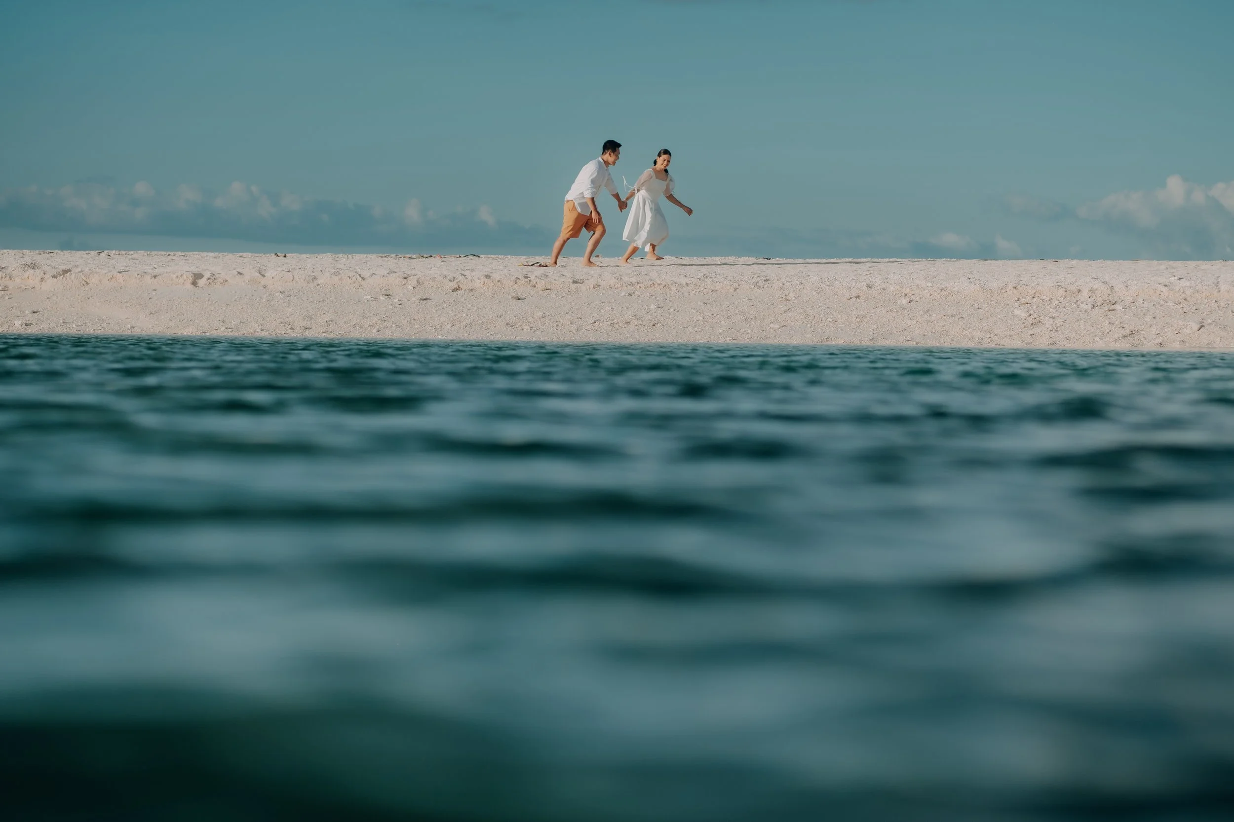 A couple walking hand in hand on a sandy beach, with the ocean in the foreground and a clear sky in the background.