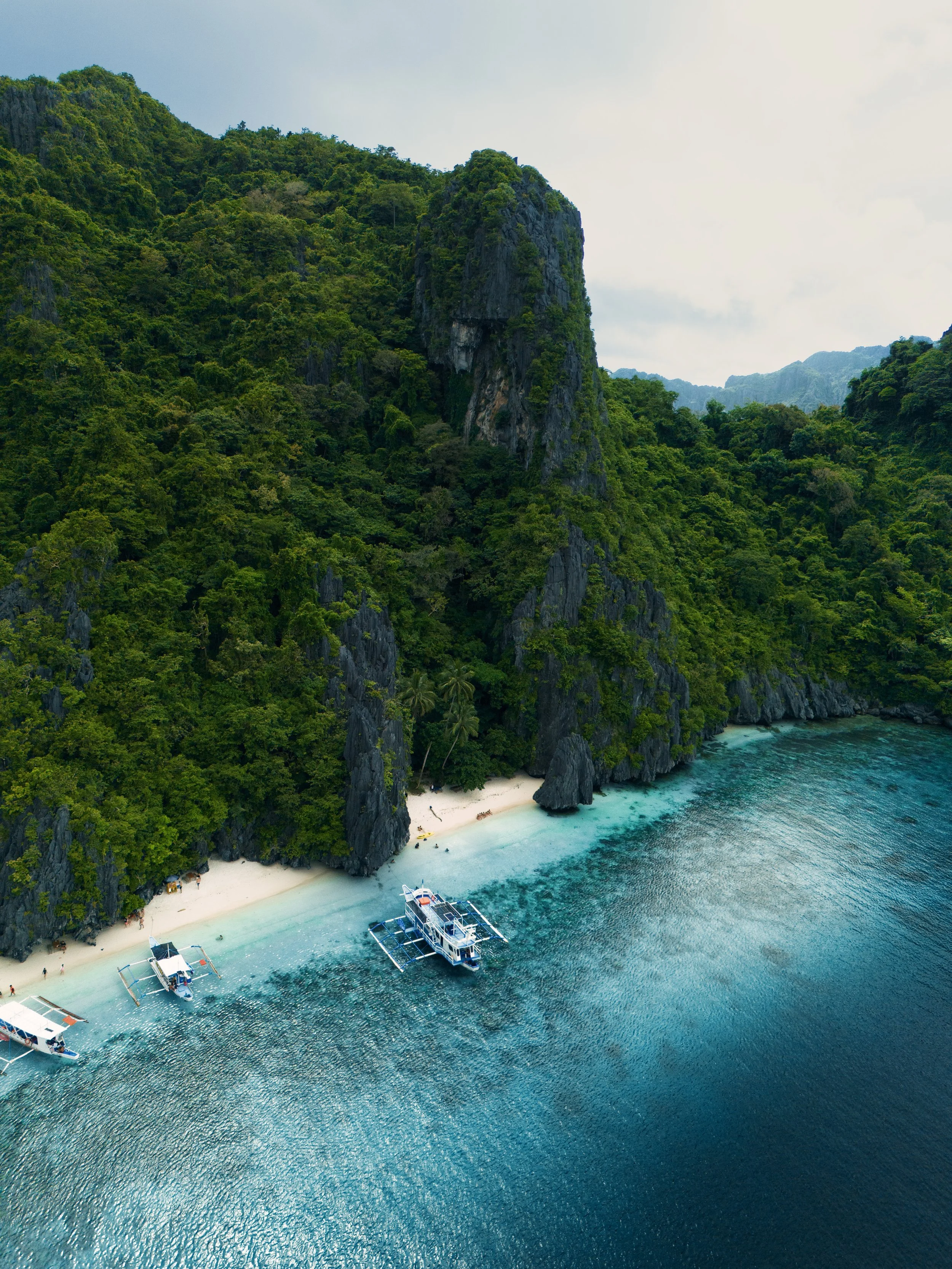 Aerial view of a tropical beach with white sand, green foliage, steep cliffs, and turquoise waters, with boats anchored near the shore.