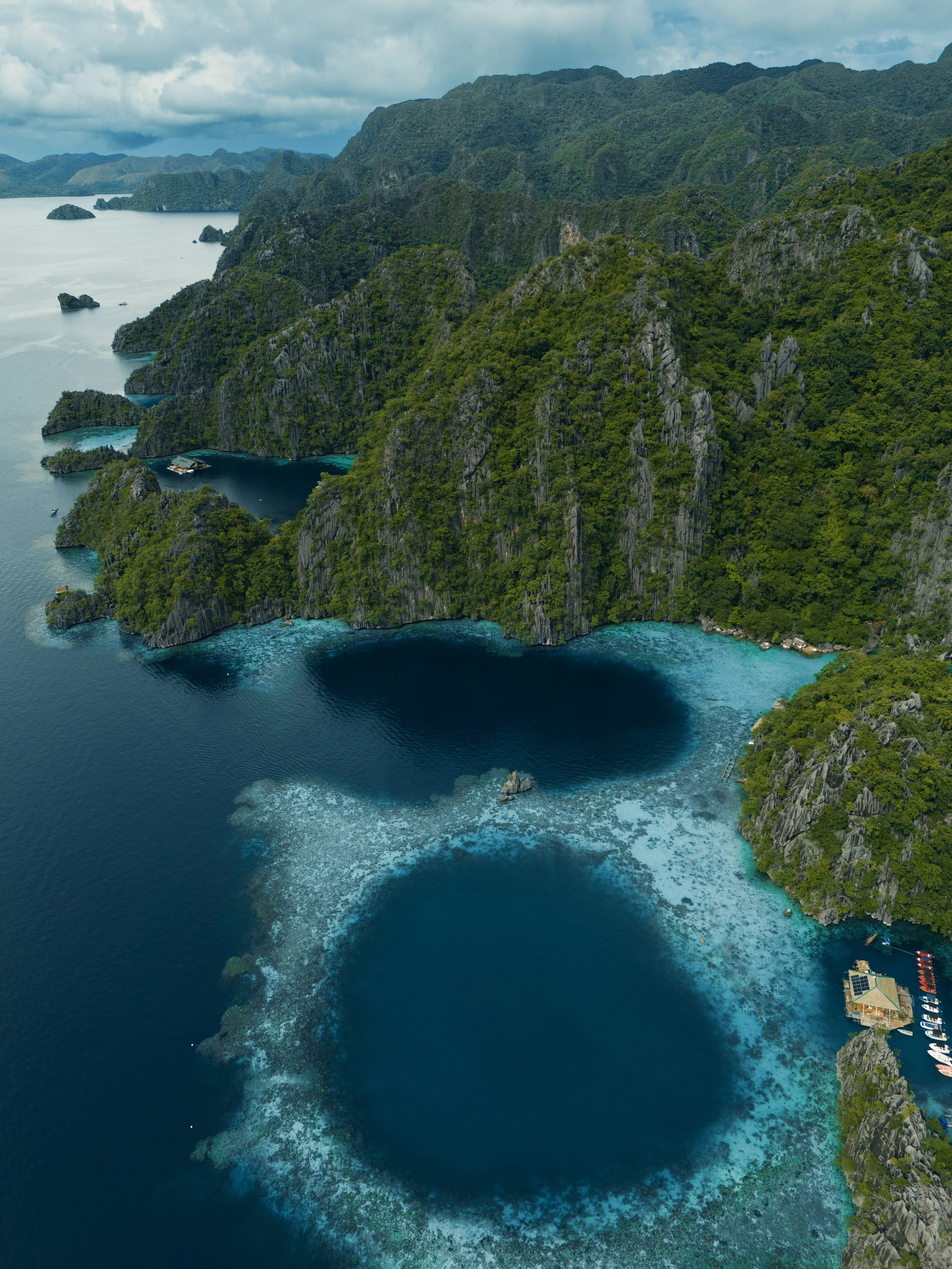 Aerial view of a tropical island with steep green cliffs and a large circular oceanic hole or sinkhole in the water surrounded by a coral reef.
