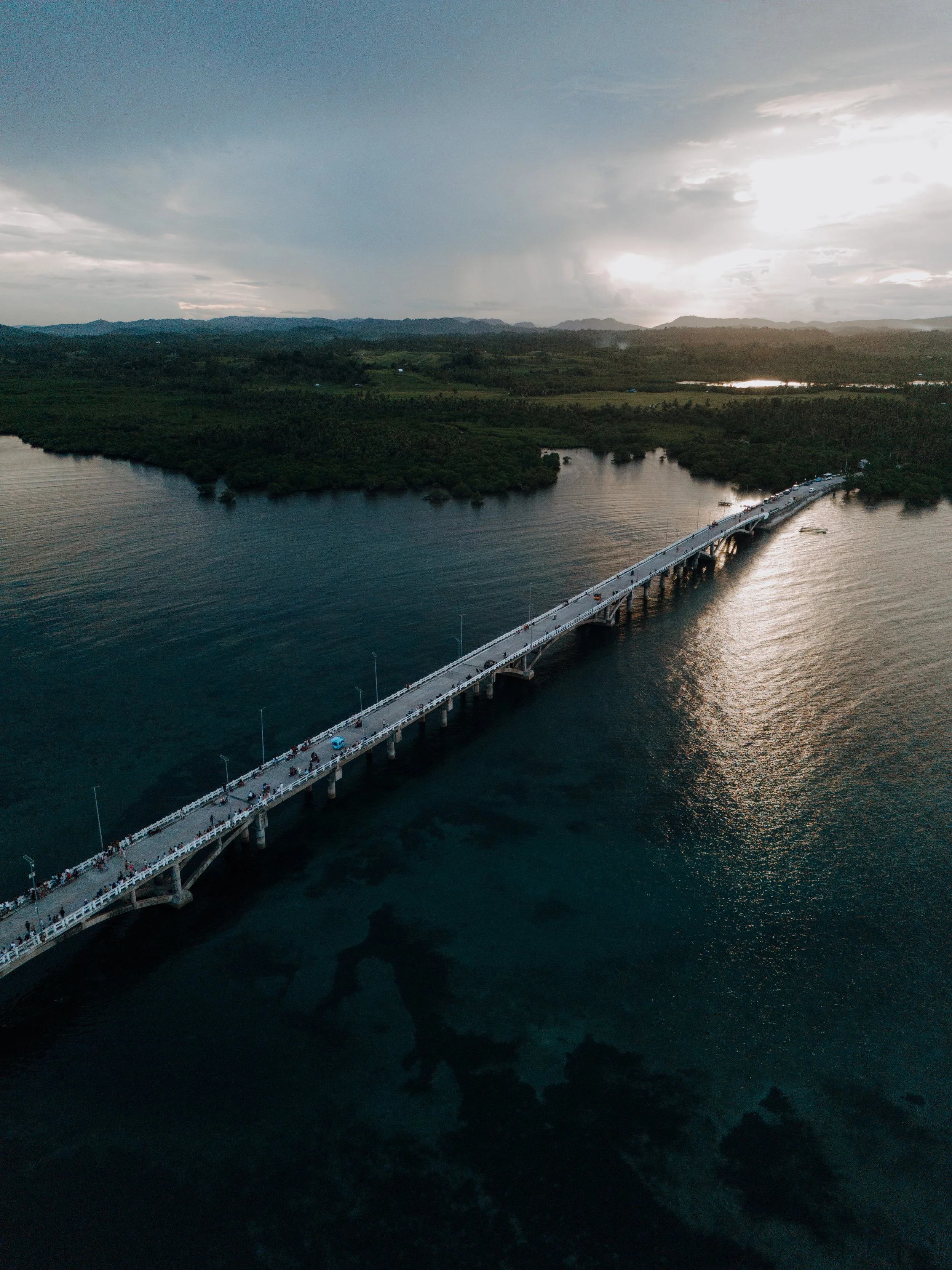 Aerial view of a long bridge extending over a body of water with green land and mountains in the background, under a cloudy sky with some sunlight breaking through.